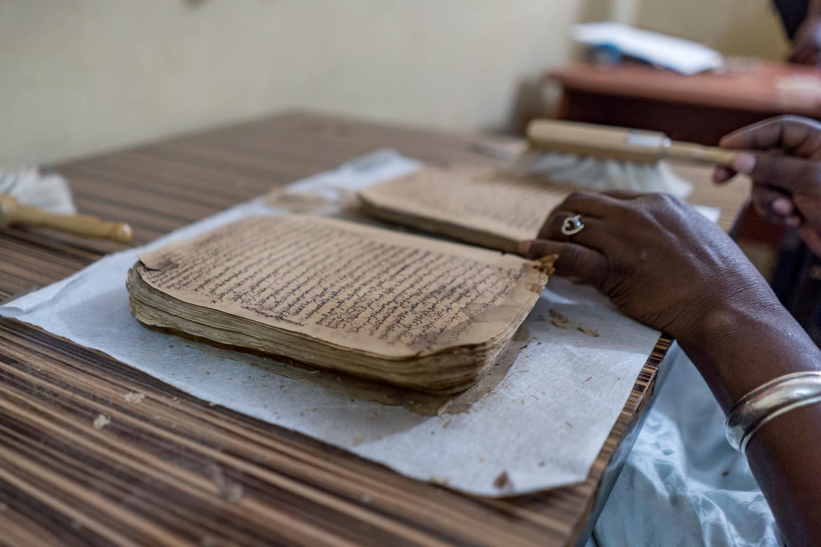Two dark hands wearing a ring and a silver bracelet hold a soft brush over a stack of brittle, yellowed manuscript leaves covered in dense lines of Arabic script, laid on white paper on a wooden table; flakes of old paper lie around them.