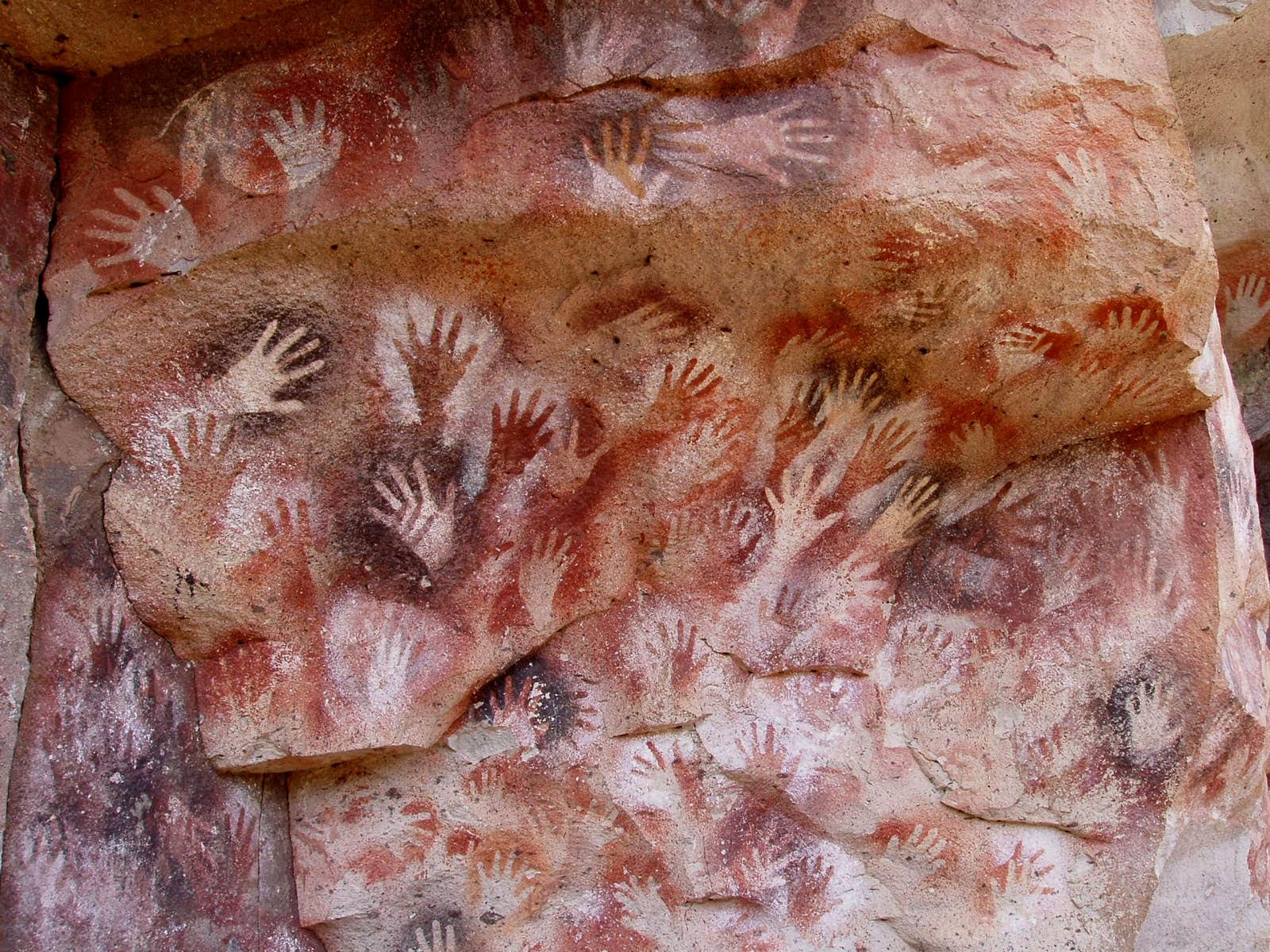 A reddish rock wall covered with dozens of overlapping hand stencils: pale outlines of open hands, fingers spread, stand out against sprayed patches of red, orange, white and black pigment.