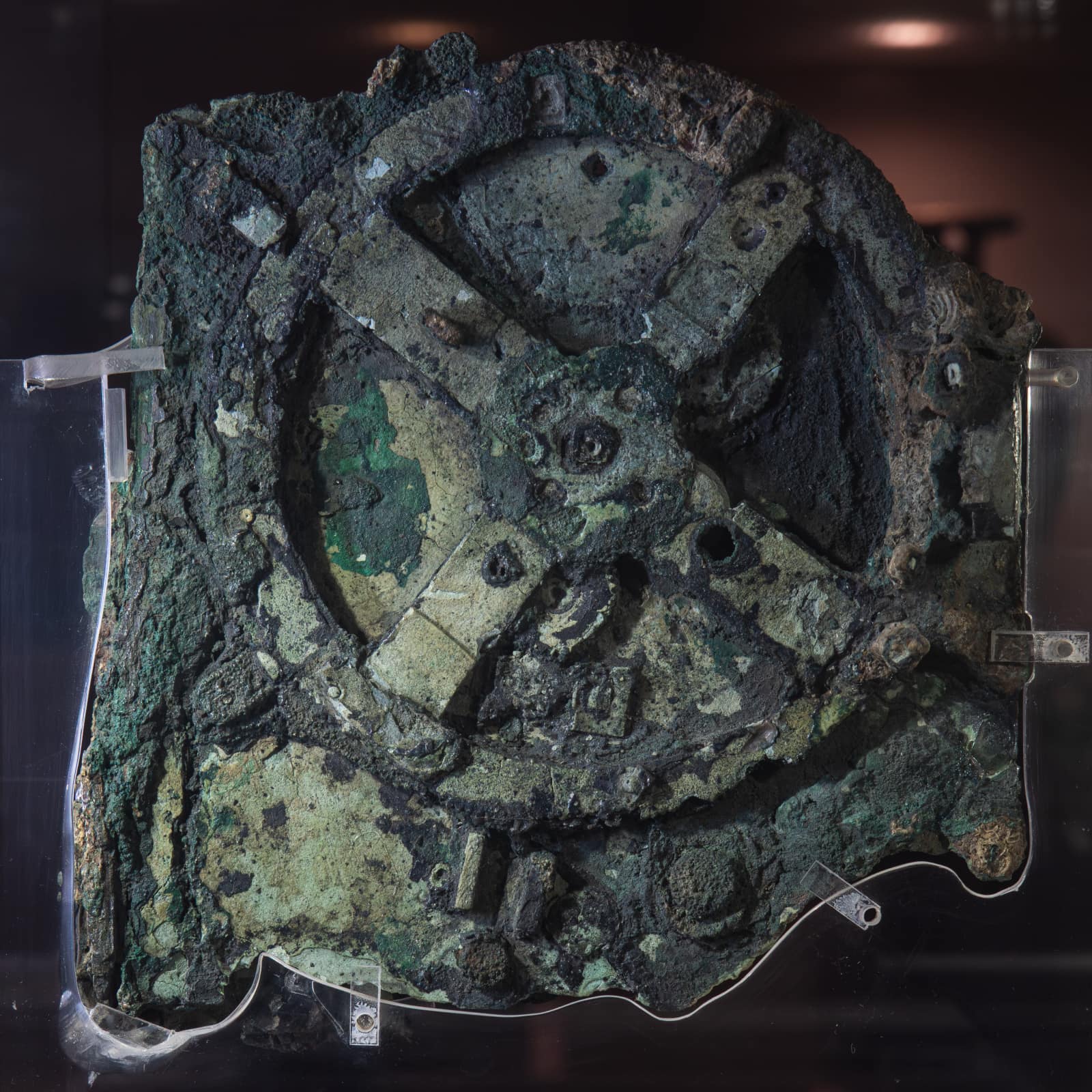 A roughly square slab of corroded bronze, green and black, held by clear mounts against a dark background: in its centre, the outline of a large gearwheel with four spokes, and around it traces of smaller wheels fused into the mass.