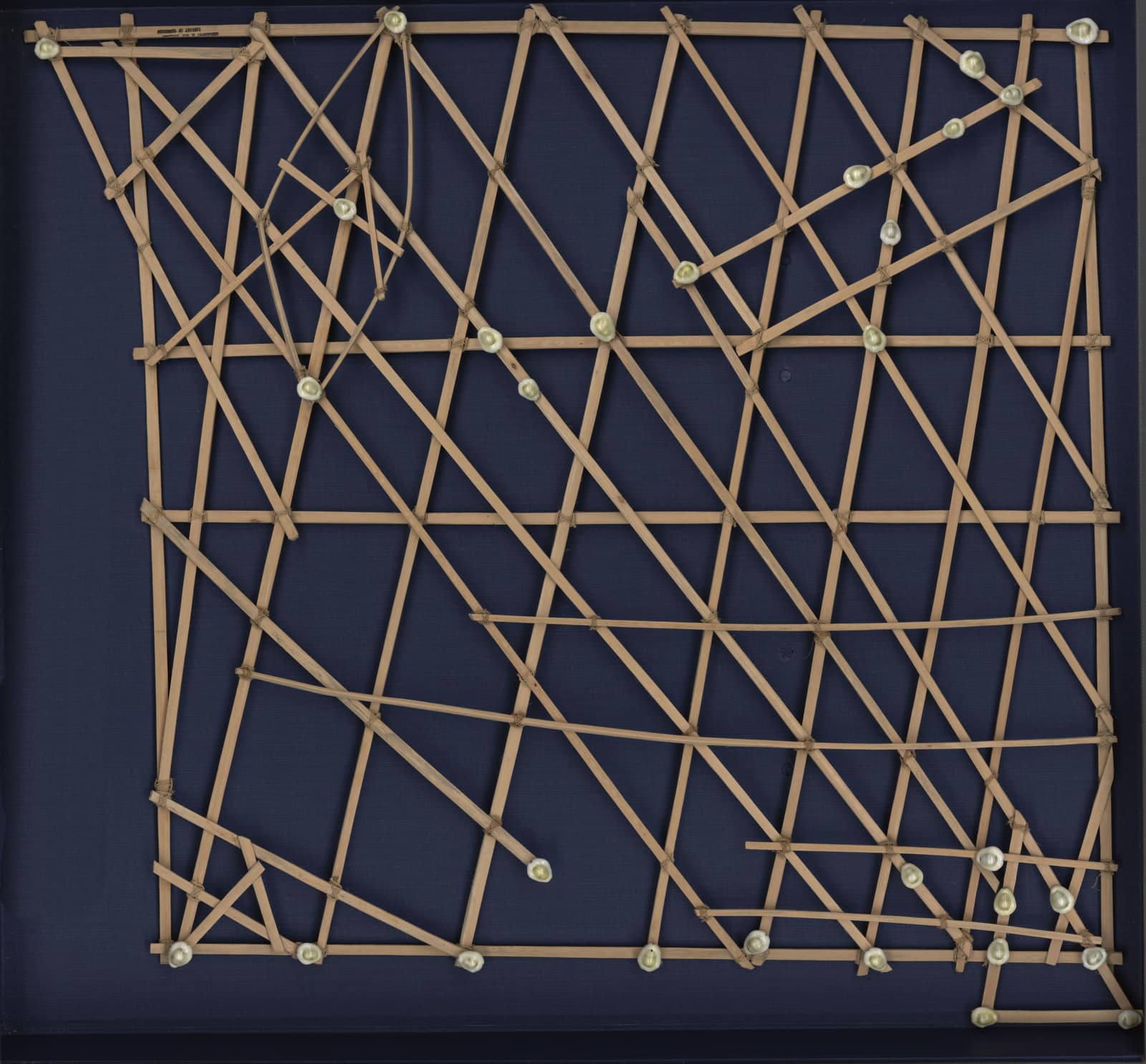 A square lattice of thin wooden sticks lashed together with fibre on a dark blue backing: straight sticks cross diagonally, a few curved sticks sweep across the frame, and small white shells are tied at many of the crossings.