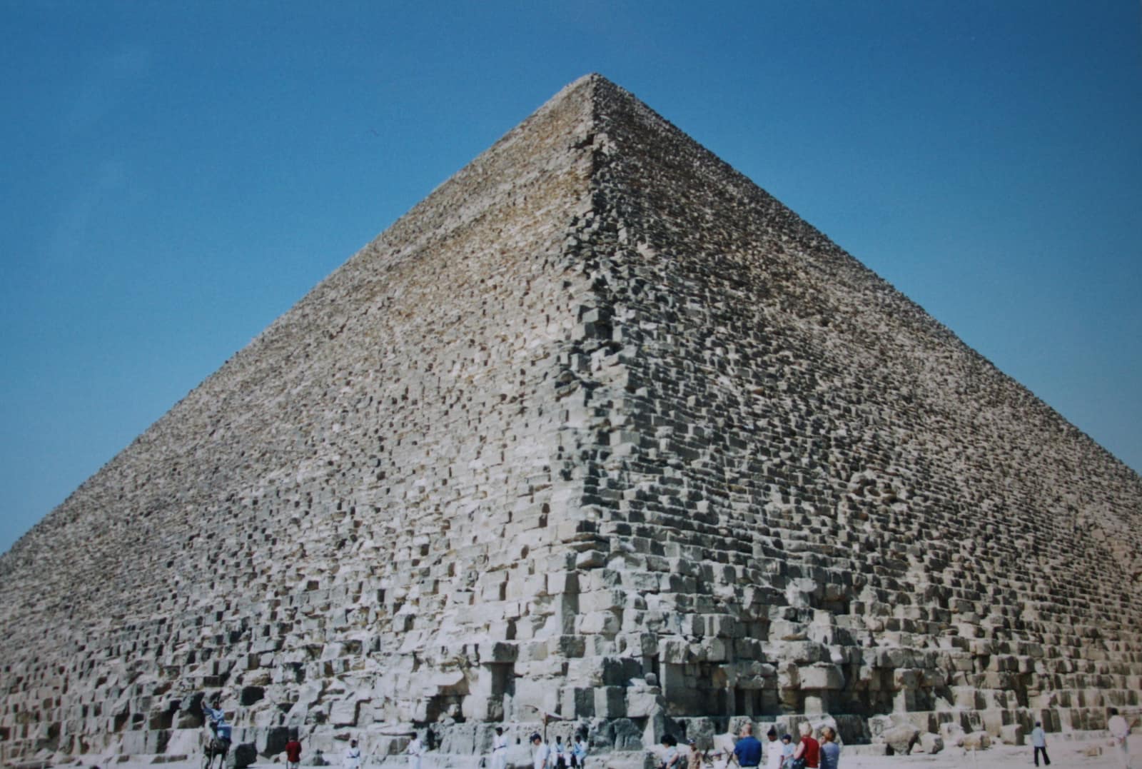 The Great Pyramid seen from close to one corner against a deep blue sky: its stepped faces of weathered, sand coloured limestone blocks rise to a point, with a scatter of tiny visitors along the base giving the scale.
