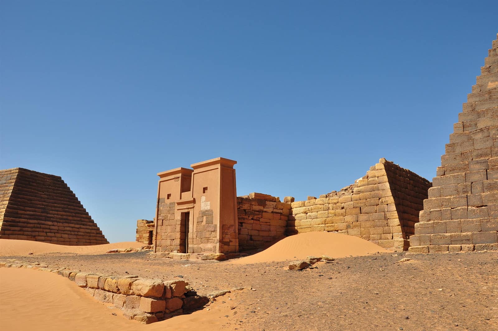 In pale orange sand under a clear blue sky, a small sandstone chapel with a gateway of two sloping towers stands among the remains of steep pyramids: on the right, a tall pyramid of stepped courses; behind and to the left, lower pyramids cut short, their tops gone.