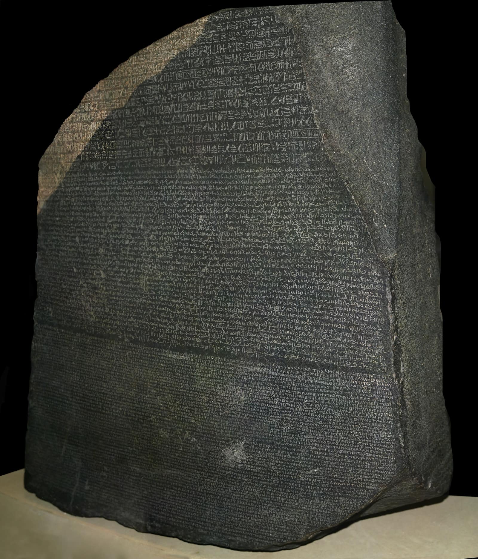 The Rosetta Stone lit against a black background: a dark grey granodiorite slab with a broken, sloping top edge and a band of pinkish stone at its upper left corner, its face covered from edge to edge in rows of small incised writing, in three bands that are hard to tell apart at this distance.