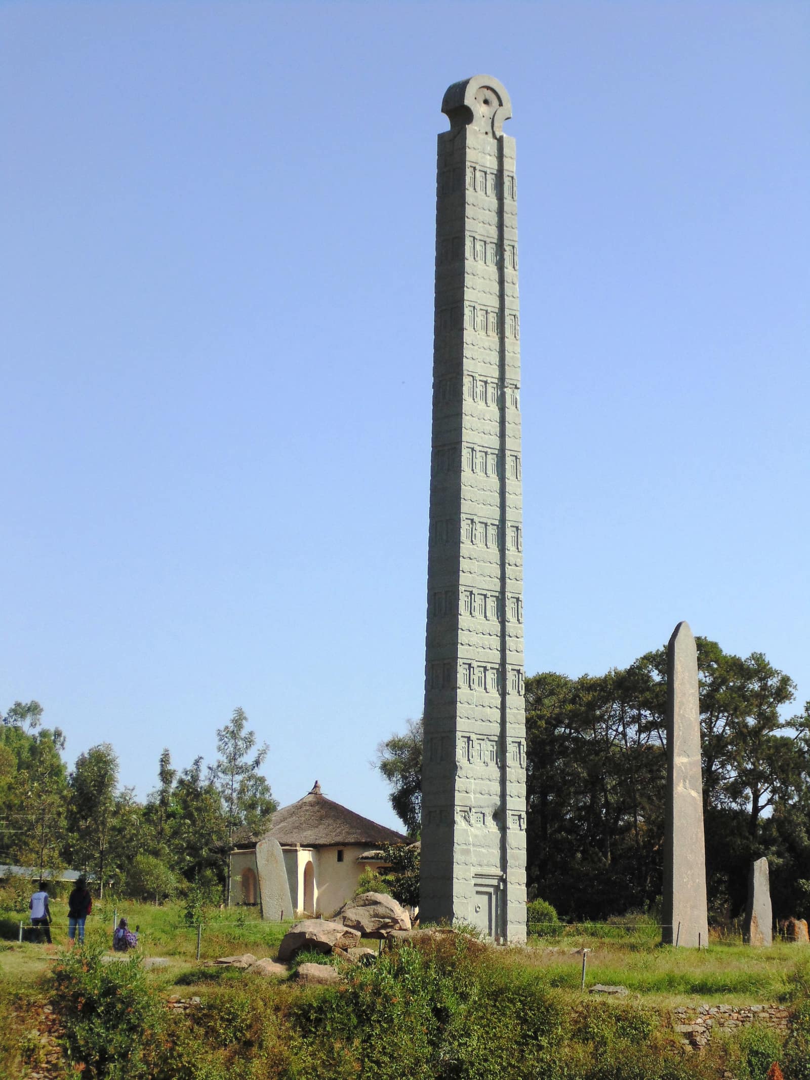 A very tall, slender grey granite stela standing in a green park under a blue sky, carved on its face with rows of false windows and beams like the storeys of a tower and topped by a rounded head; a smaller plain stela stands to its right and a round thatched building sits at its foot among trees.