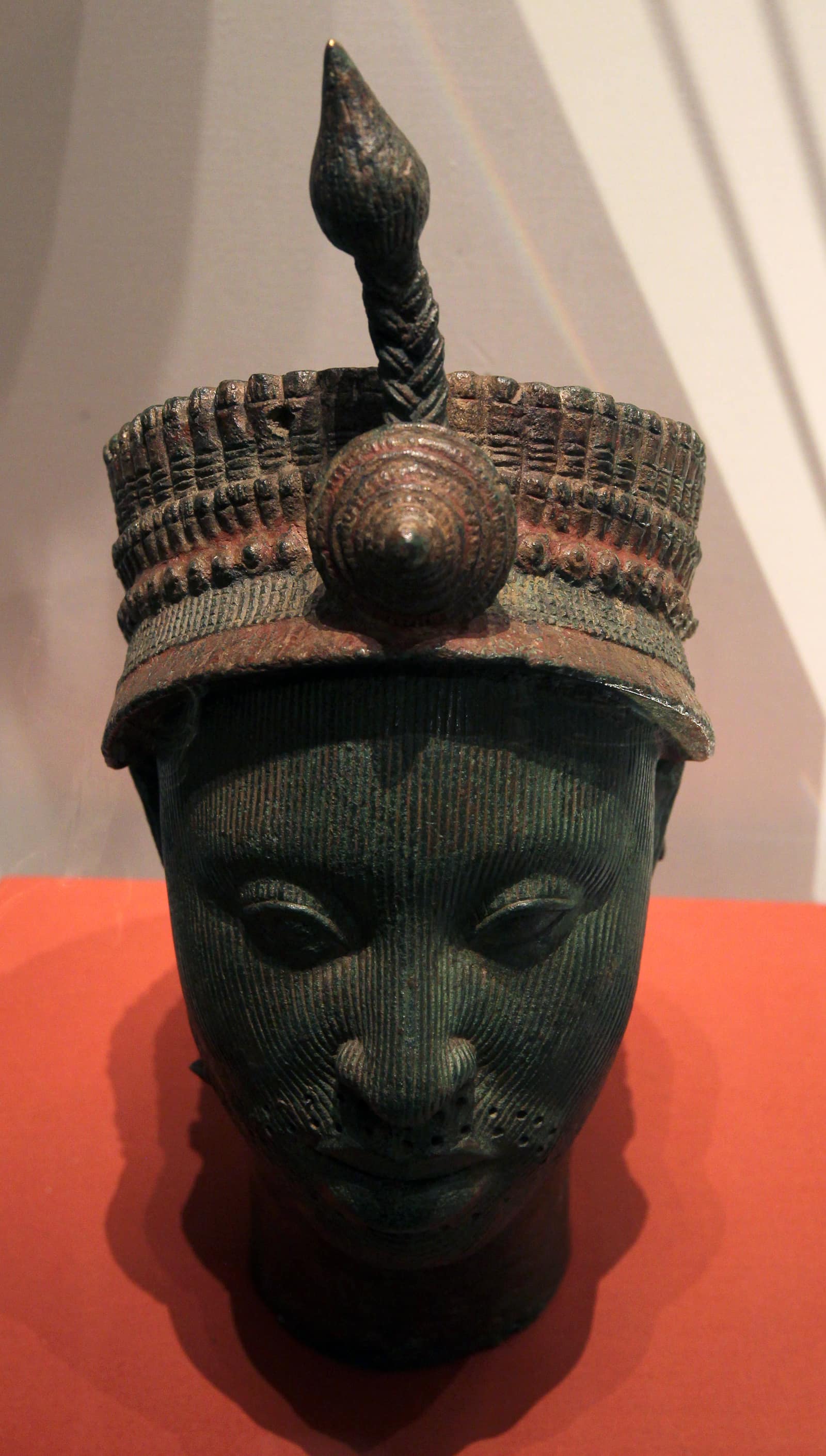 A life-like cast metal head with a dark green patina, shown against a red and white museum wall: the calm face has full lips, almond eyes and fine parallel lines running down the whole face; it wears a tall beaded crown with a rosette at the front and a plume-like crest rising from the top.