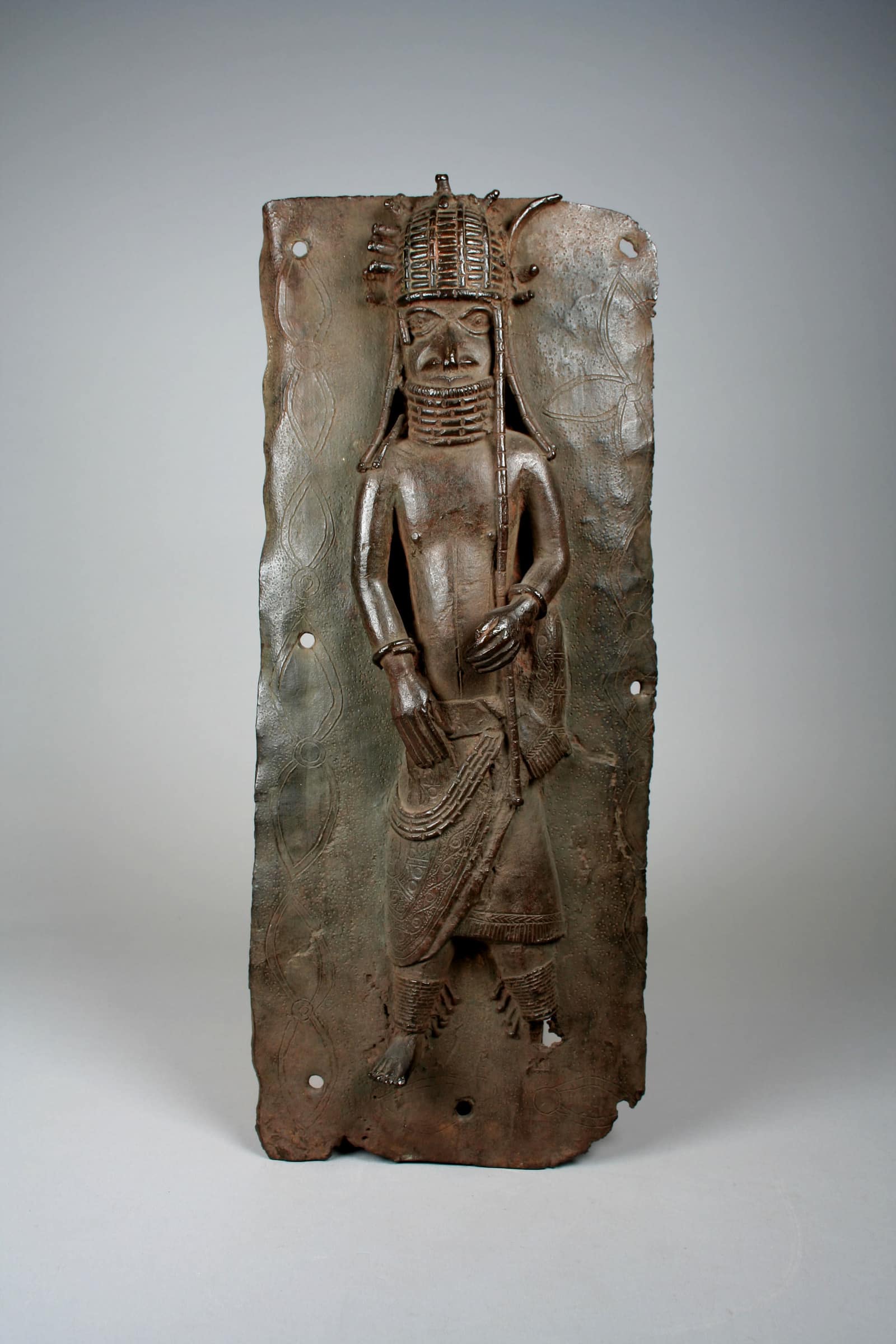 A tall rectangular brass plaque, dark brown with age and pierced with nail holes at its edges, photographed against a grey background: a single standing man in high relief faces forward, wearing a tall beaded cap with projecting spikes, a high collar of beads up to his chin, a wrapped skirt with patterned edges and beaded bands at the ankles, holding a long staff; faint engraved leaf and quatrefoil patterns cover the background.