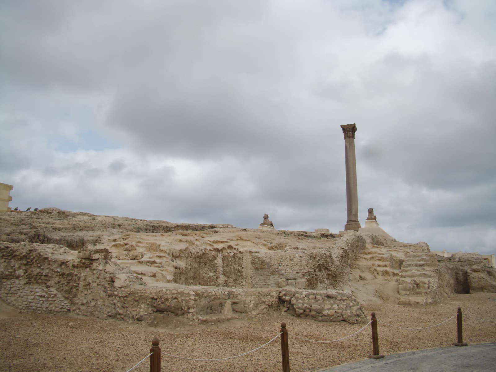 Low ruined walls and foundations of pale limestone on a rise under a grey, clouded sky at the site of the Serapeum in Alexandria, with a single tall granite column standing alone on the right and a rope barrier along the path in the foreground.