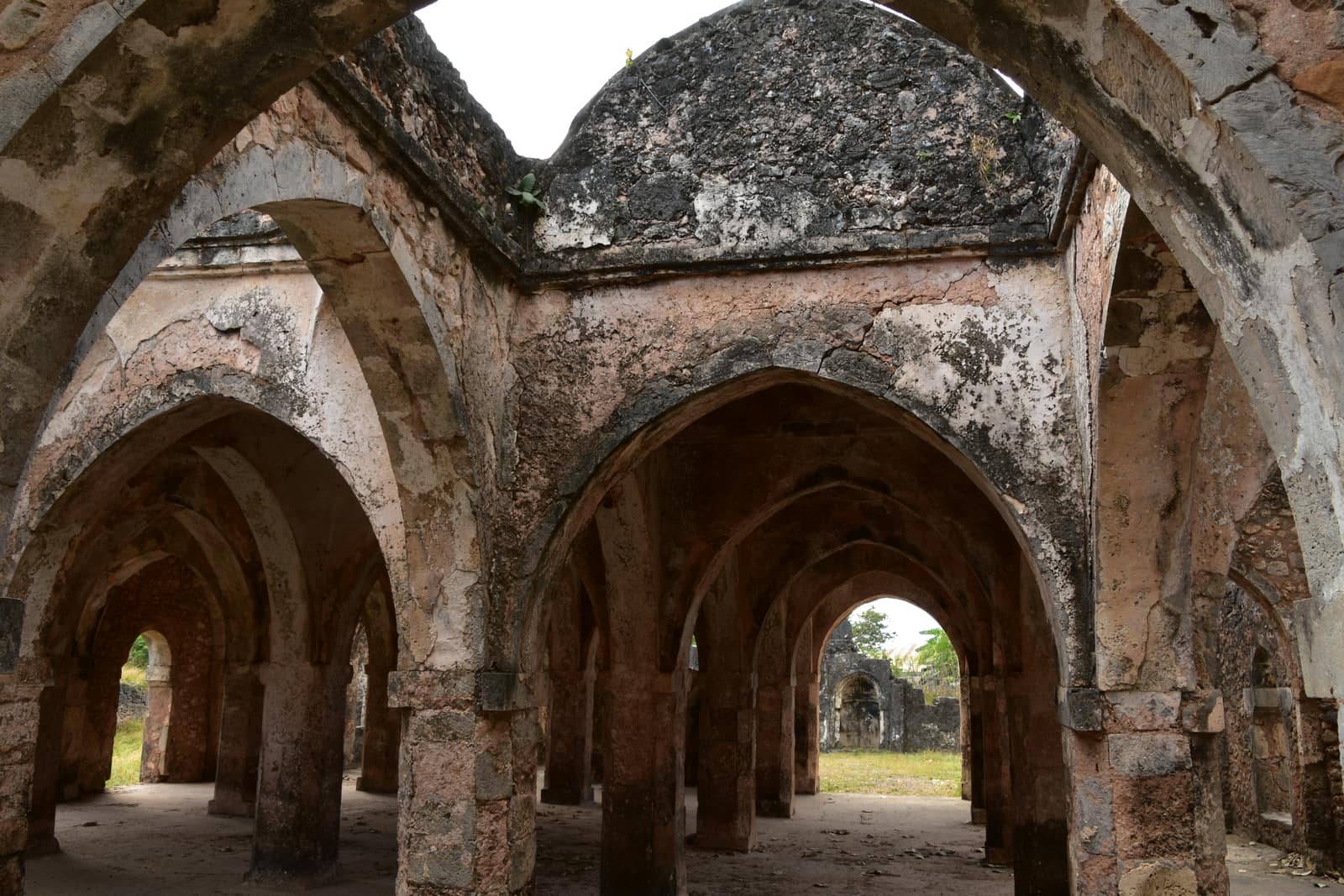 Inside the ruined Great Mosque of Kilwa: rows of weathered grey and ochre coral stone arches on slender pillars carry a roof of low domes, some patched with dark lichen, with daylight and green trees showing through the far arches.