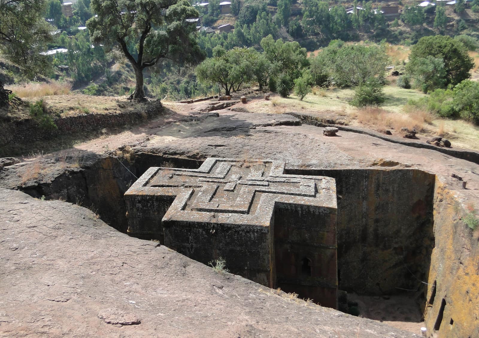 Bete Giyorgis seen from the rim of its pit: a church carved down into reddish rock, its flat roof shaped as a large cross of three stepped outlines, standing free inside a deep square trench, with bare rock, green trees and hillsides of the town around it.