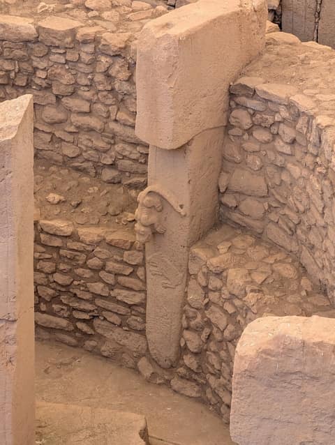 Inside a round enclosure of rough stone walls, a tall T-shaped limestone pillar with a heavy rectangular head; on its shaft a predator is carved in high relief, head and forelegs jutting out as if climbing down, above the flat outline of a wild boar.