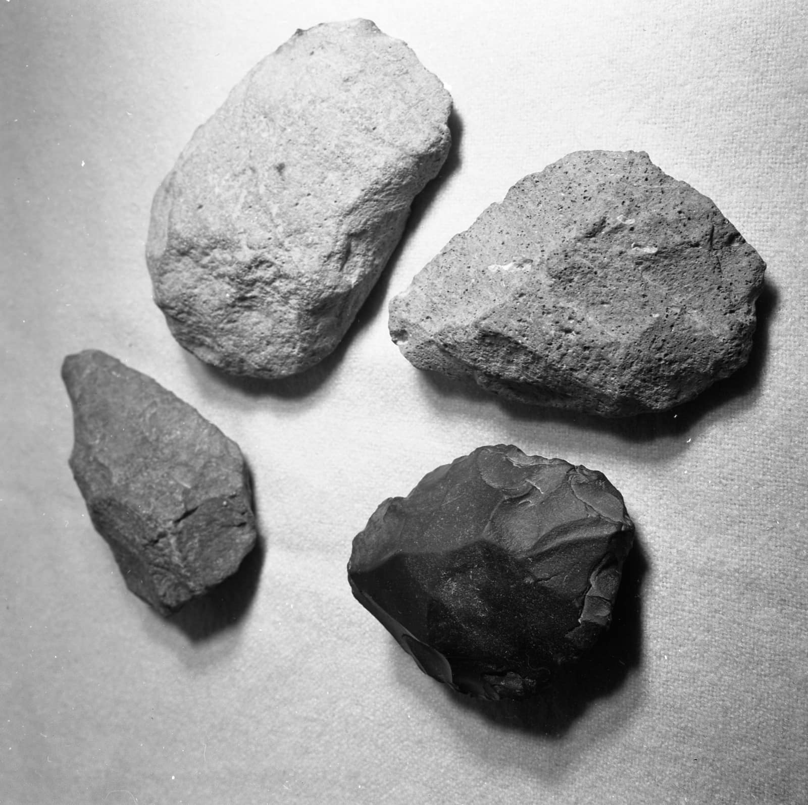 Black and white photograph from 1959: four roughly worked stones lie on a pale cloth, two light and pitted, one smaller and grey, one dark with sharp flake scars, recorded among archaeological finds from Gesher Benot Ya'aqov.