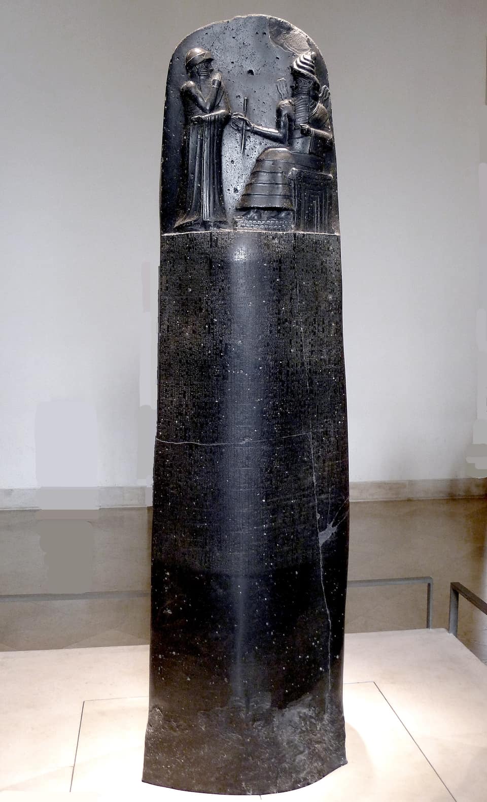 A tall black basalt stele, rounded at the top, standing in a pale gallery. In the relief at its summit a bearded king stands with one hand raised before a seated god who holds out a rod and a ring; below, the whole polished shaft is covered in close columns of tiny cuneiform signs.