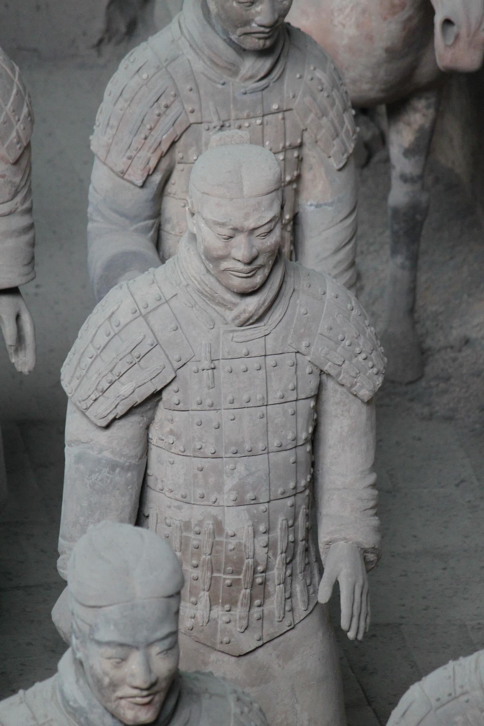 Seen from slightly above, a life-size grey clay soldier in Pit 1 wears armour of small riveted plates over a knee-length robe; his hair is tied in a topknot, his eyes are lowered, his empty right hand once held a weapon. Other soldiers and the leg of a clay horse stand around him.