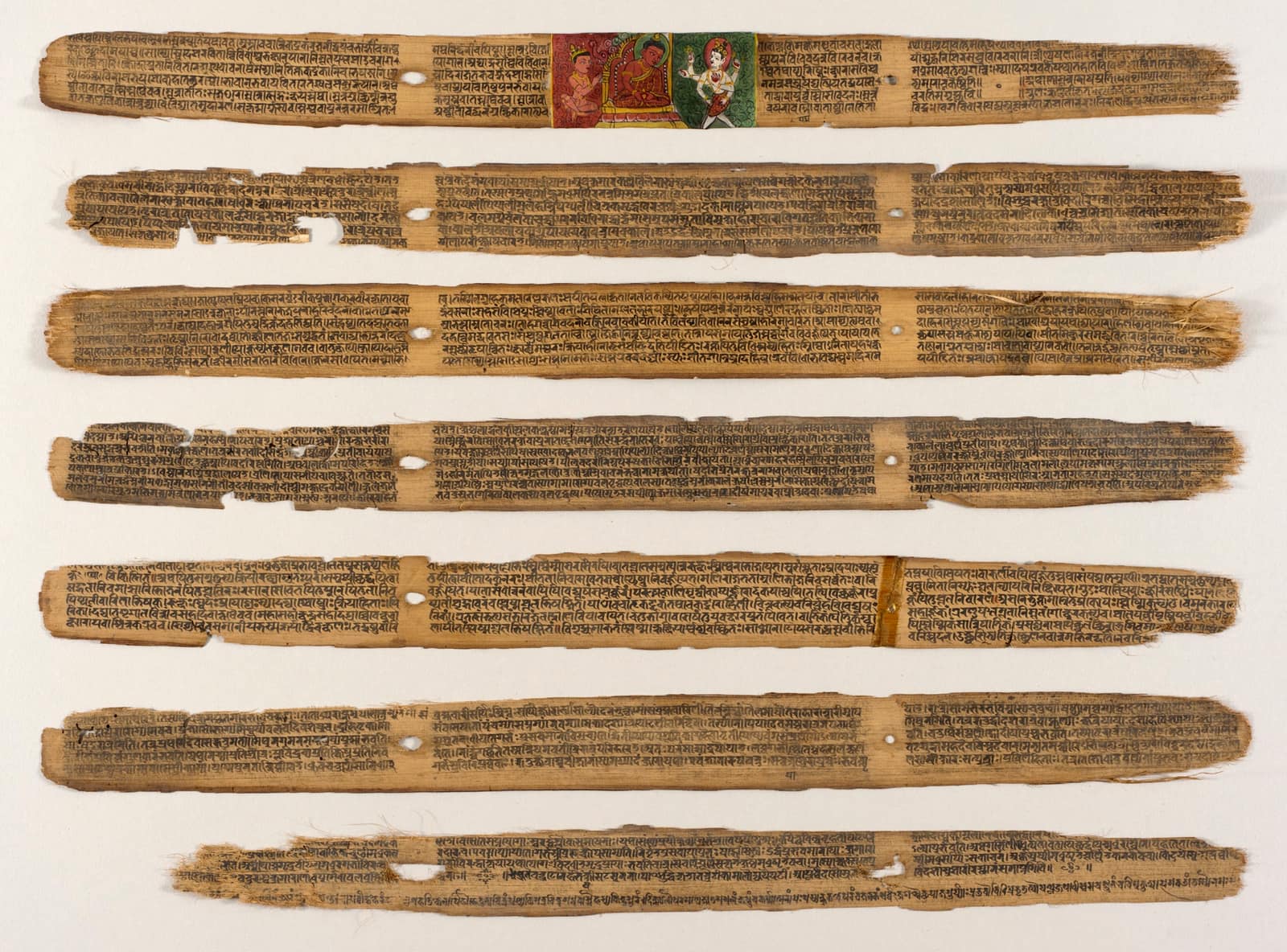 Seven long, narrow palm leaves laid one above the other on a white ground, their brown edges frayed and pierced by string holes, each covered in fine lines of dark Sanskrit script; on the top leaf a small painted panel shows seated figures in red, green and gold.