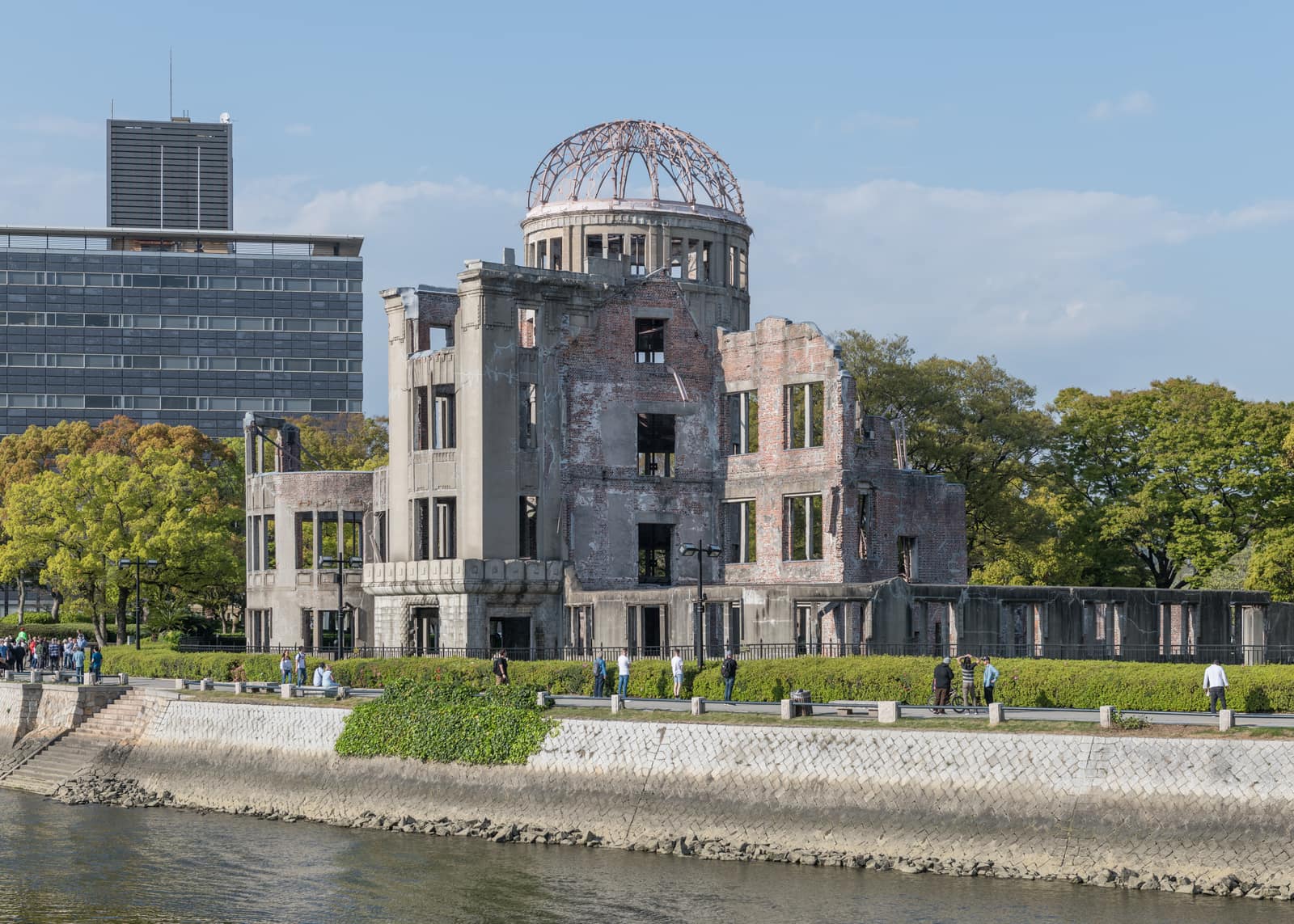 Seen across a river on a clear spring day, the ruin of a brick and stone building with empty window openings and broken upper walls, crowned by the bare steel ribs of a dome; green trees and a modern office block stand behind, and people walk along the embankment in front.