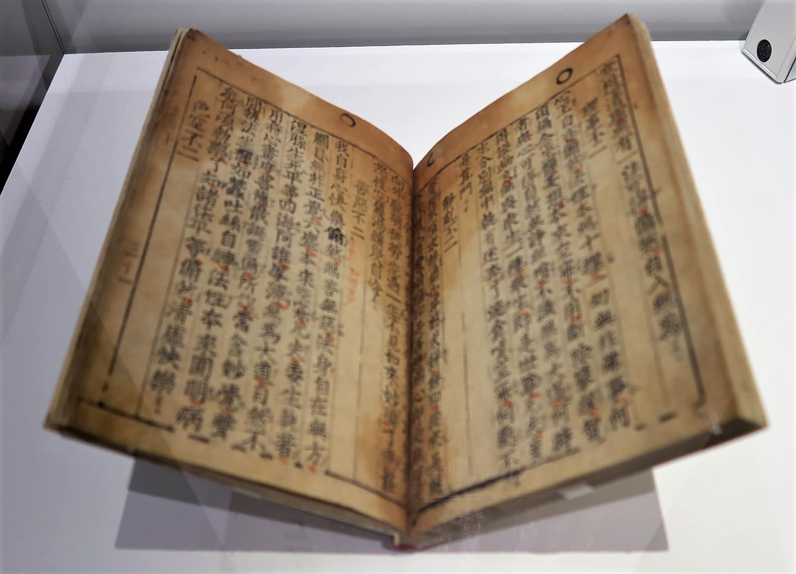 An old book of yellowed paper lying open in a glass case, its two pages printed in columns of black Chinese characters inside thin ruled frames, some characters slightly uneven in size and inking, with small red marks added by hand beside the text.