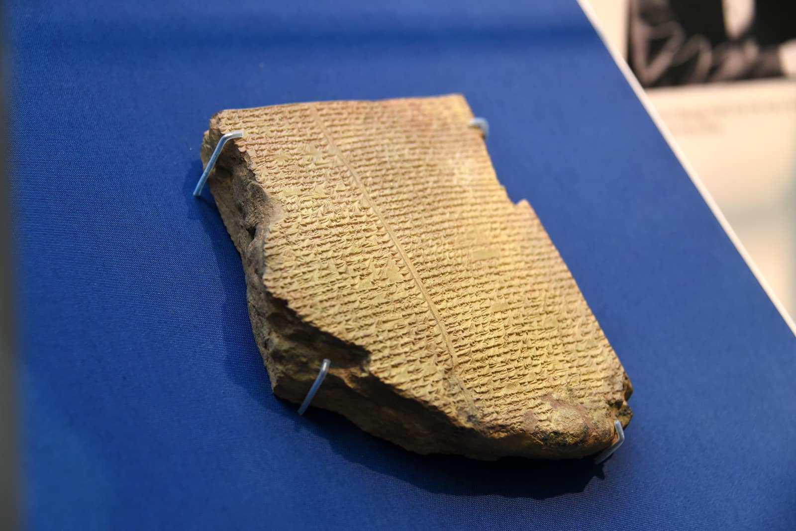 A broken slab of baked clay, the colour of sand, lying on blue cloth in a museum case and held by small metal clips; its surface is packed with rows of tiny wedge-shaped cuneiform signs in two columns divided by a ruled line, and its left and lower edges are broken away.