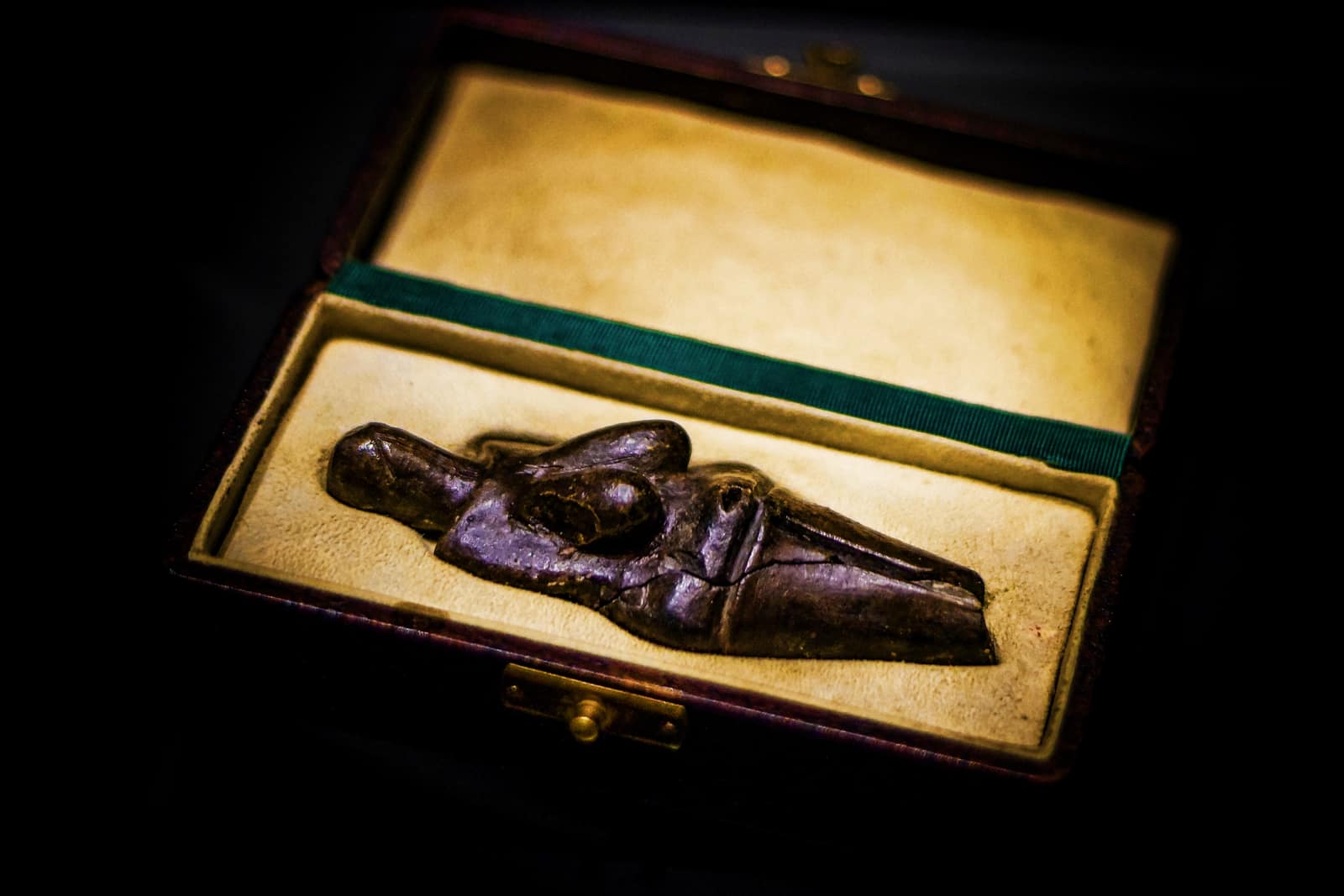 A small dark figure of fired clay lying in an open box lined with pale fabric: a woman with heavy breasts and wide hips, a faceless head with slits for eyes, and legs that end in a point, lit against a black background.