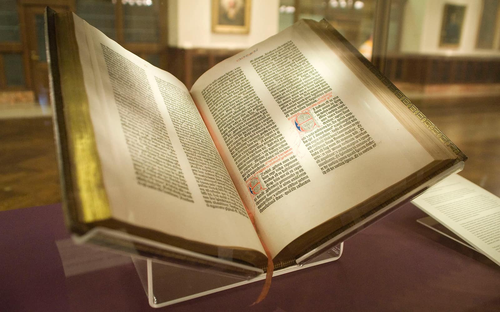 A large open book on a clear display stand in a dim library room: two pages of dense black Latin type set in two columns each, with a red and blue initial and a few red headings, the thick paper edges and leather binding catching the light.