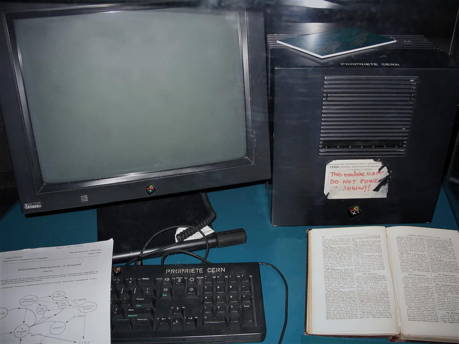 A black NeXT cube computer in a glass display case beside its monitor and keyboard; a white label on the cube reads, in red handwriting, This machine is a server, DO NOT POWER IT DOWN; in front lie an open book and a printed sheet with a hand drawn diagram.