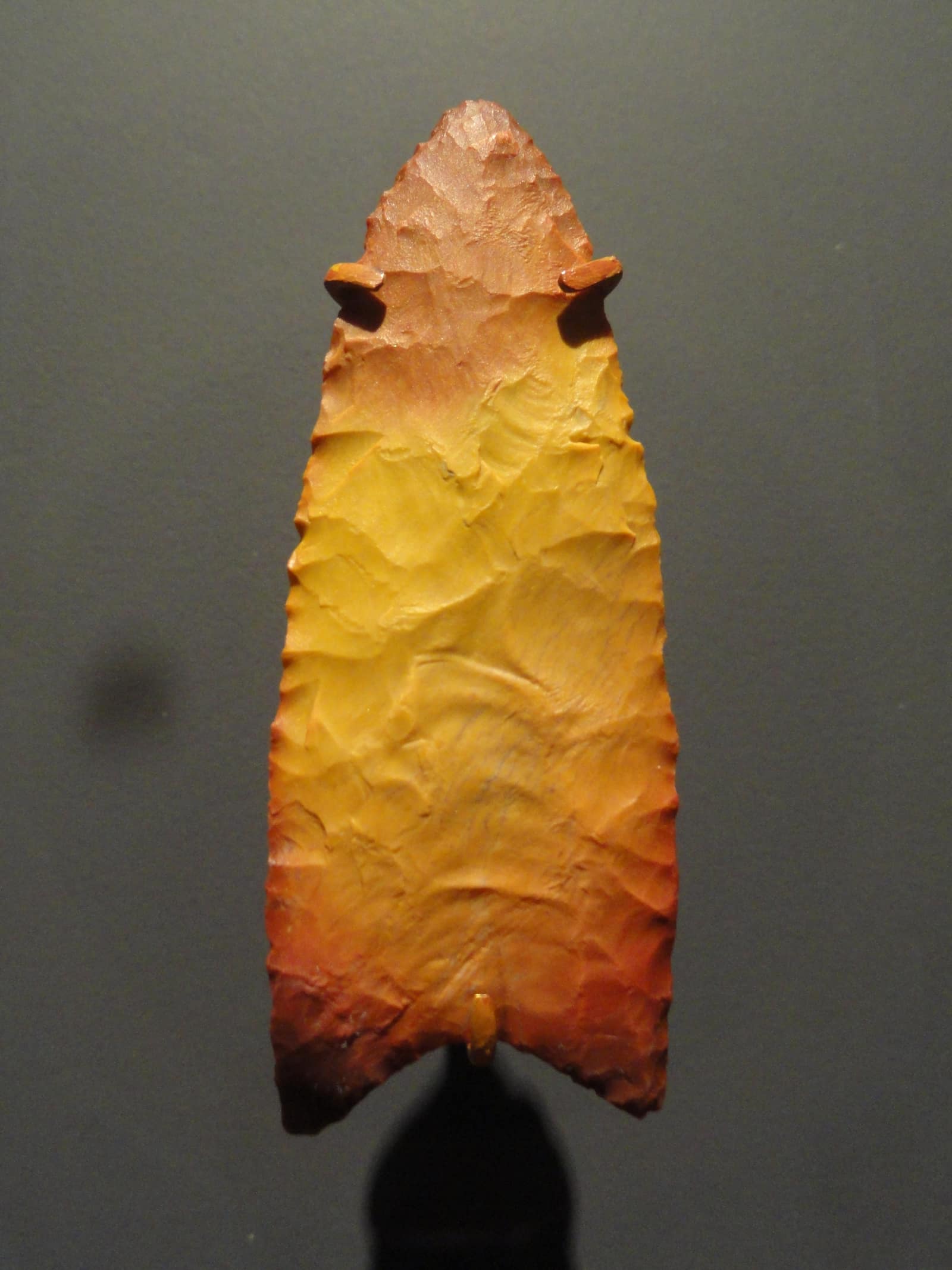 A single stone spear point, long and leaf shaped, standing upright against a dark grey background: yellow chert shading to orange and red at the tip and base, its faces covered with shallow scars where flakes were struck off, its edges finely serrated and its base concave, with a shallow channel running up from the base.