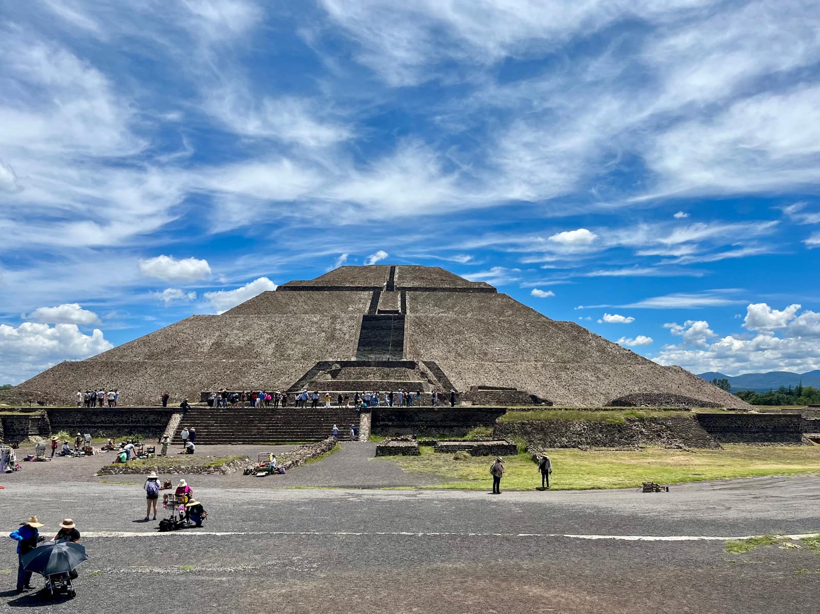 A vast stepped pyramid of grey stone seen from the front under a blue sky streaked with white cloud: a central staircase rises through its terraces to a flat top where small figures of visitors stand; in the foreground, a paved plaza, low platforms and a few people with sun hats and umbrellas.