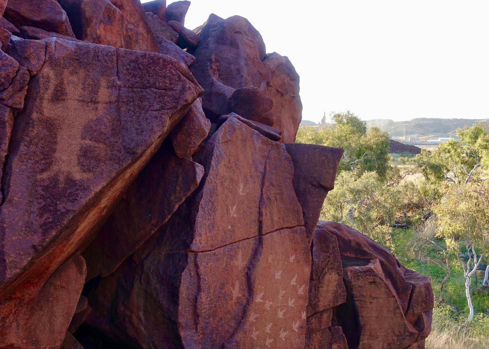 A pile of dark red boulders in low sunlight; on the flat face of one, rows of small pale three-pronged marks have been pecked through the dark surface into the lighter stone. To the right, beyond green trees, the chimneys and buildings of an industrial plant stand on the horizon.