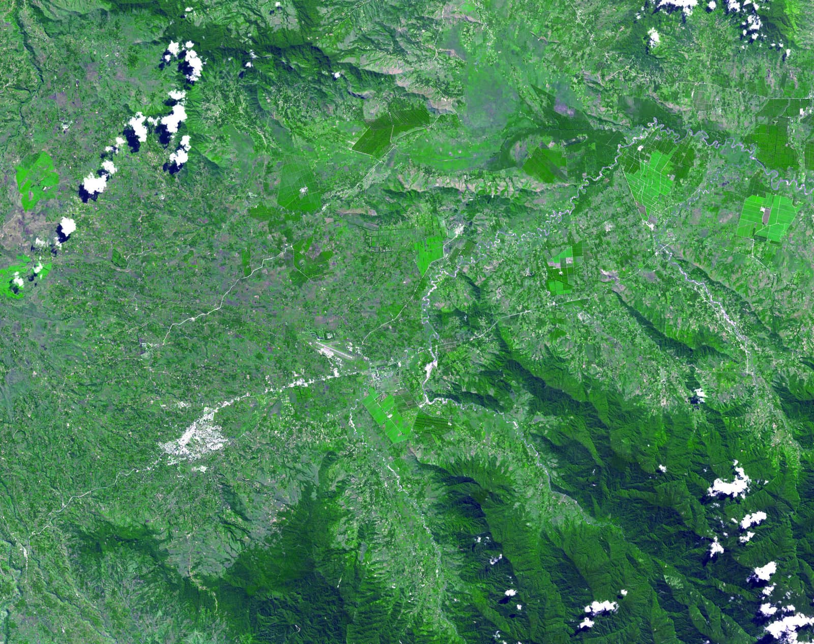 A satellite view of a green highland valley in false colour: forested ridges and folded mountains frame a broad valley floor patterned with small fields, bright rectangular plantations, winding rivers, a pale patch of town and an airstrip, with a few white clouds and their shadows at the edges.
