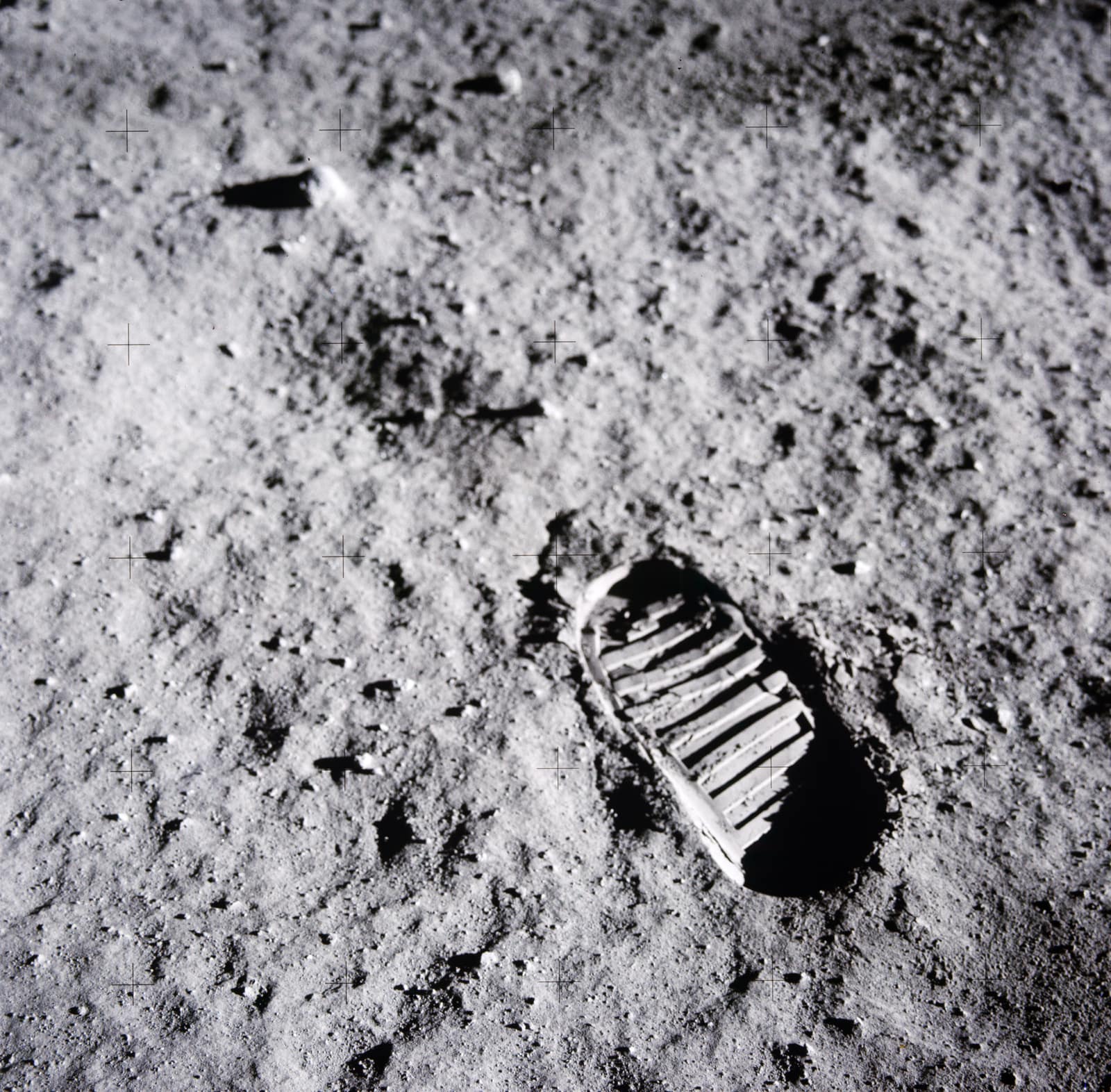 Black and white close view of grey lunar soil, fine as powder and scattered with small stones: in the lower right, a single boot print with deep parallel ridges from the sole, its edges sharp; small black crosses from the camera's reference grid are spaced evenly across the image.