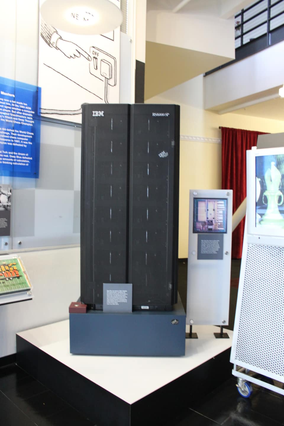 A tall black computer cabinet in two panels on a dark blue plinth in a museum hall, marked IBM at the top left and RS/6000 SP at the top right, with a small label in front; behind it, a panel with a drawing of a hand pressing an off switch, and to the right a screen showing a chess piece.