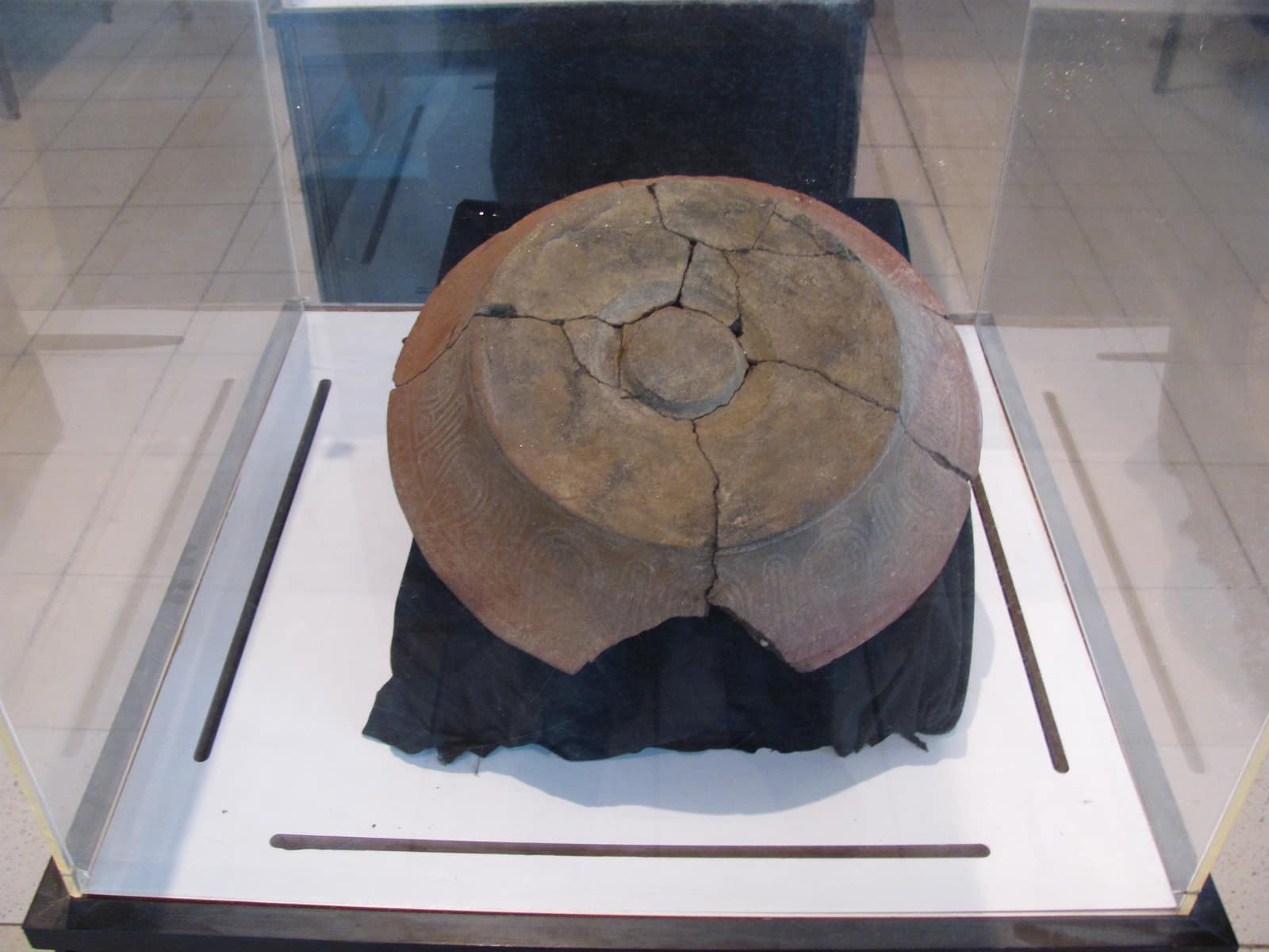 A wide earthenware dish, reddish brown, shown upside down in a glass case on a dark cloth: its flat base and a central foot ring are visible, the body is cracked and glued from many pieces, and around the sloping sides runs faint stamped decoration of fine dotted lines in curving bands.