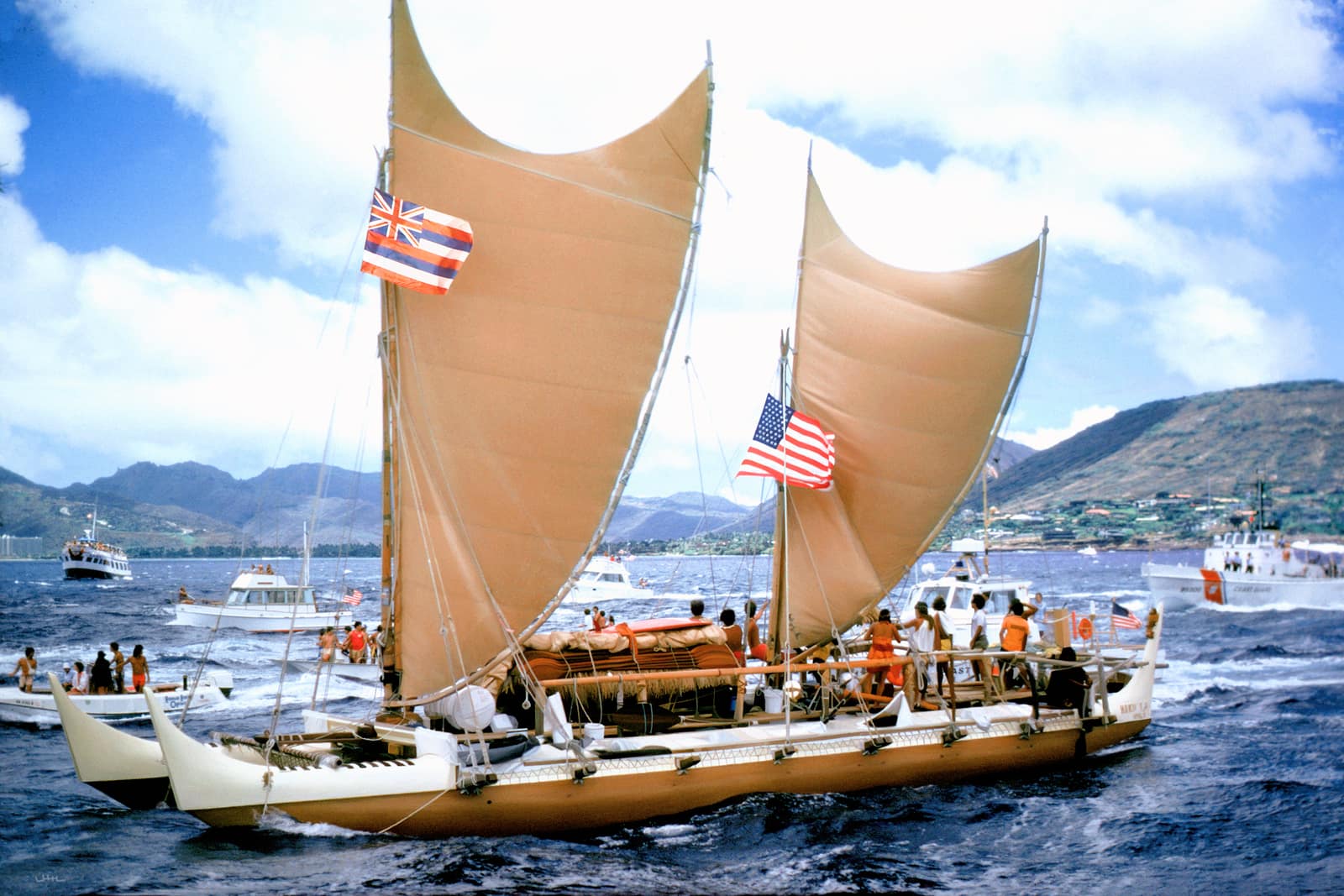 A double-hulled voyaging canoe under two tan crab claw sails crosses choppy blue water towards the shore, flying the Hawaiian flag and the United States flag; crew in bright clothes stand on the deck between the hulls, small motorboats follow, and green mountains rise behind under white clouds.