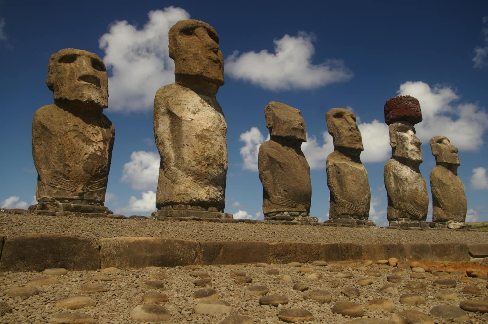 Six weathered stone figures stand in a row on a long stone platform under a deep blue sky with white clouds: long faces with heavy brows and narrow chins, arms carved along the sides of their bodies; one wears a red stone cylinder on its head, and the ground before them is paved with rounded stones.