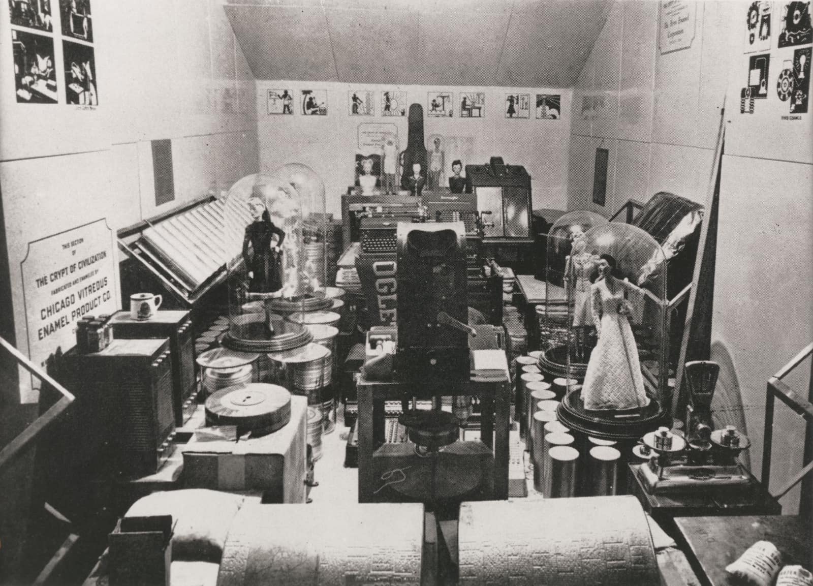 Une photographie en noir et blanc d'une pièce longue et étroite aux murs émaillés clairs et au plafond en pente, encombrée d'objets avant la fermeture : piles de cylindres métalliques et de boîtes de film, une machine à écrire, un poste de radio, une balance, et des cloches de verre abritant des poupées en robe longue ; sur le mur de gauche, un panneau indique en anglais que cette section de la crypte a été fabriquée et émaillée par la Chicago Vitreous Enamel Product Co.