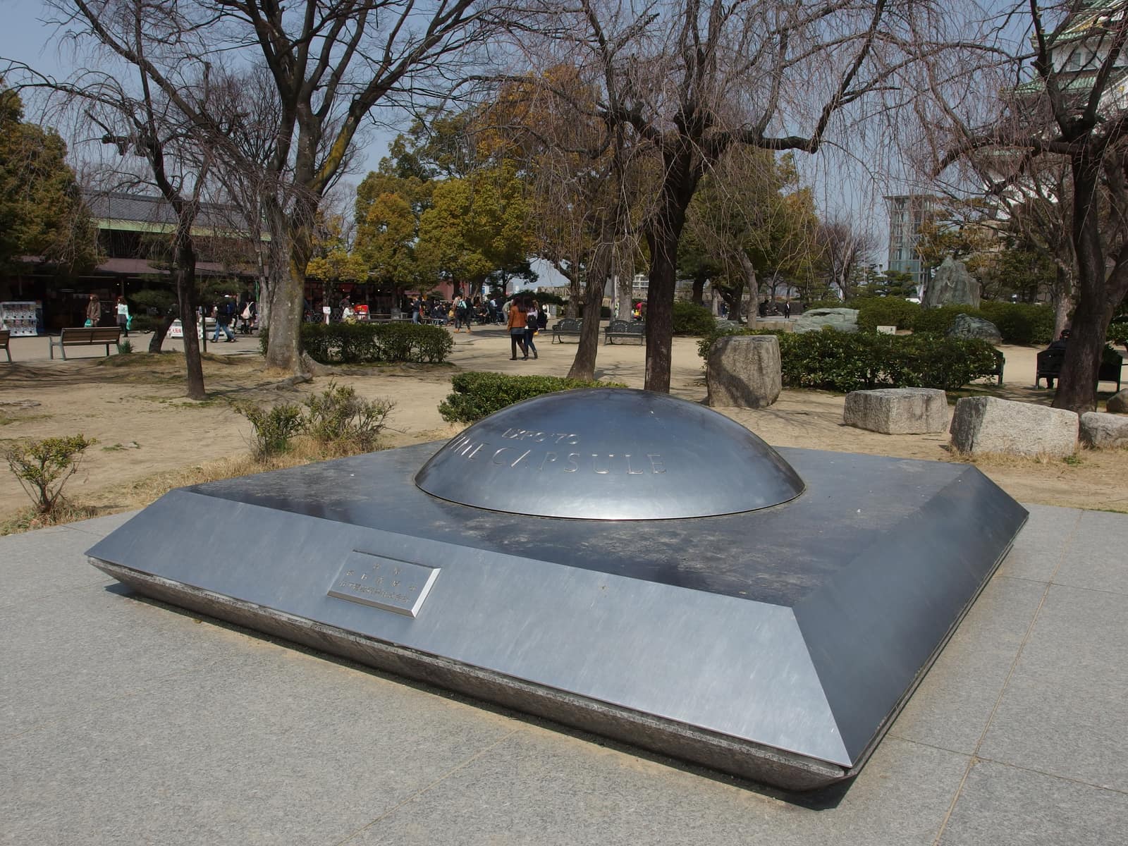 Un monument bas et carré en pierre grise polie, dans un parc : ses flancs inclinés montent vers un plateau d'où émerge un dôme d'acier brillant, comme une sphère à demi enfouie, gravé des mots Time Capsule. Derrière, des arbres d'hiver dénudés, des bancs et des promeneurs.
