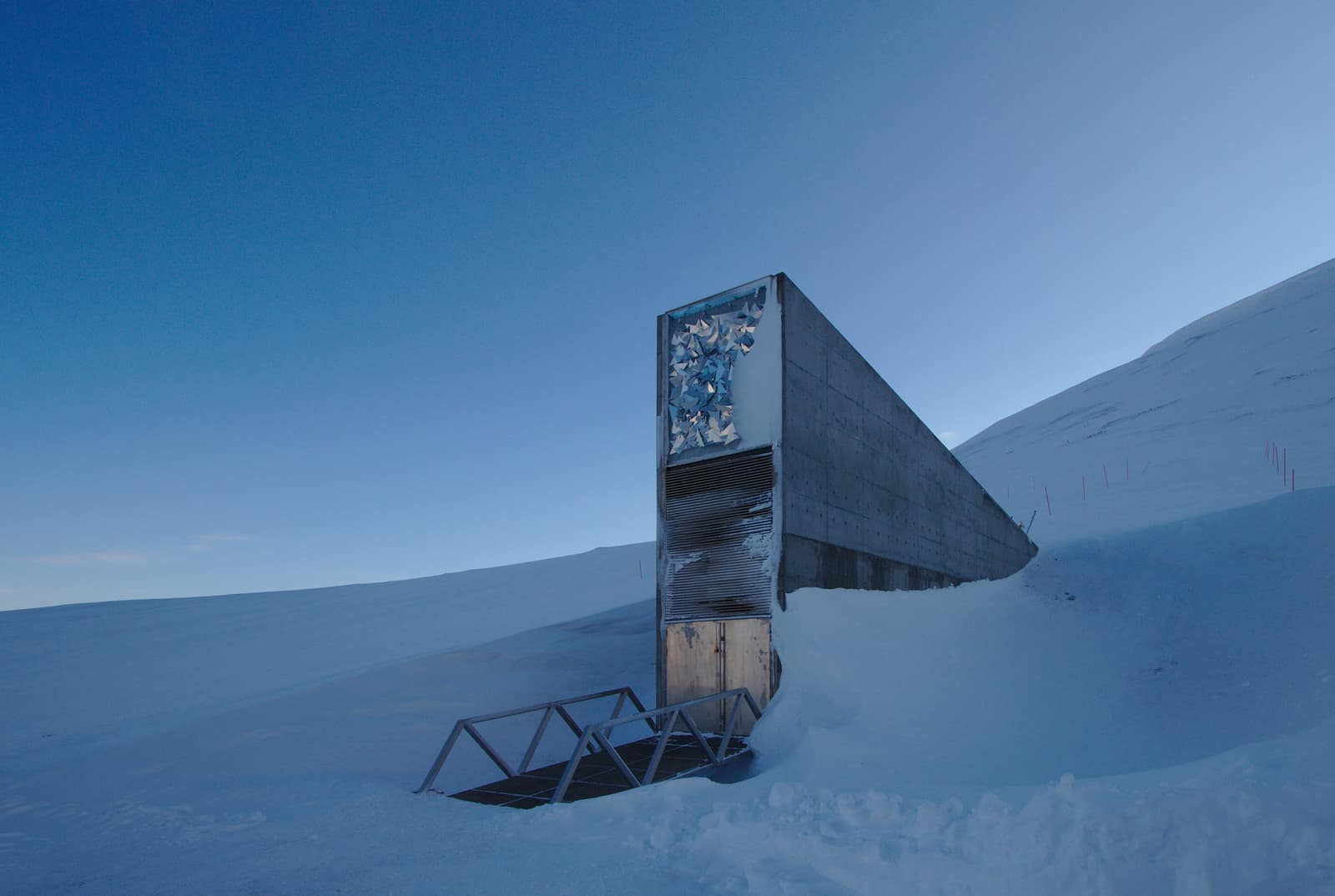 Un étroit coin de béton gris surgit d'une pente enneigée sous un ciel d'un bleu profond ; sa haute façade porte un panneau d'éclats anguleux et réfléchissants au-dessus d'une grille d'aération et d'une petite porte, à laquelle mène une passerelle métallique posée sur la neige.