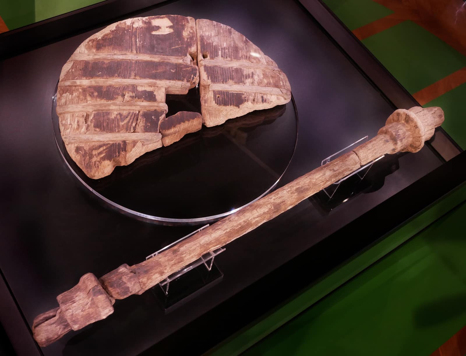 Dans une vitrine sombre, un disque de bois rond repose à plat sur un support transparent : il est fait de deux larges planches jointes bord à bord, traversées de quatre bandes plus sombres là où étaient insérées des chevilles, avec un trou carré au centre et un morceau brisé manquant à côté. Devant lui, sur de petits supports transparents, repose un long essieu de bois usé, carré aux deux extrémités, à la surface fendue et patinée.