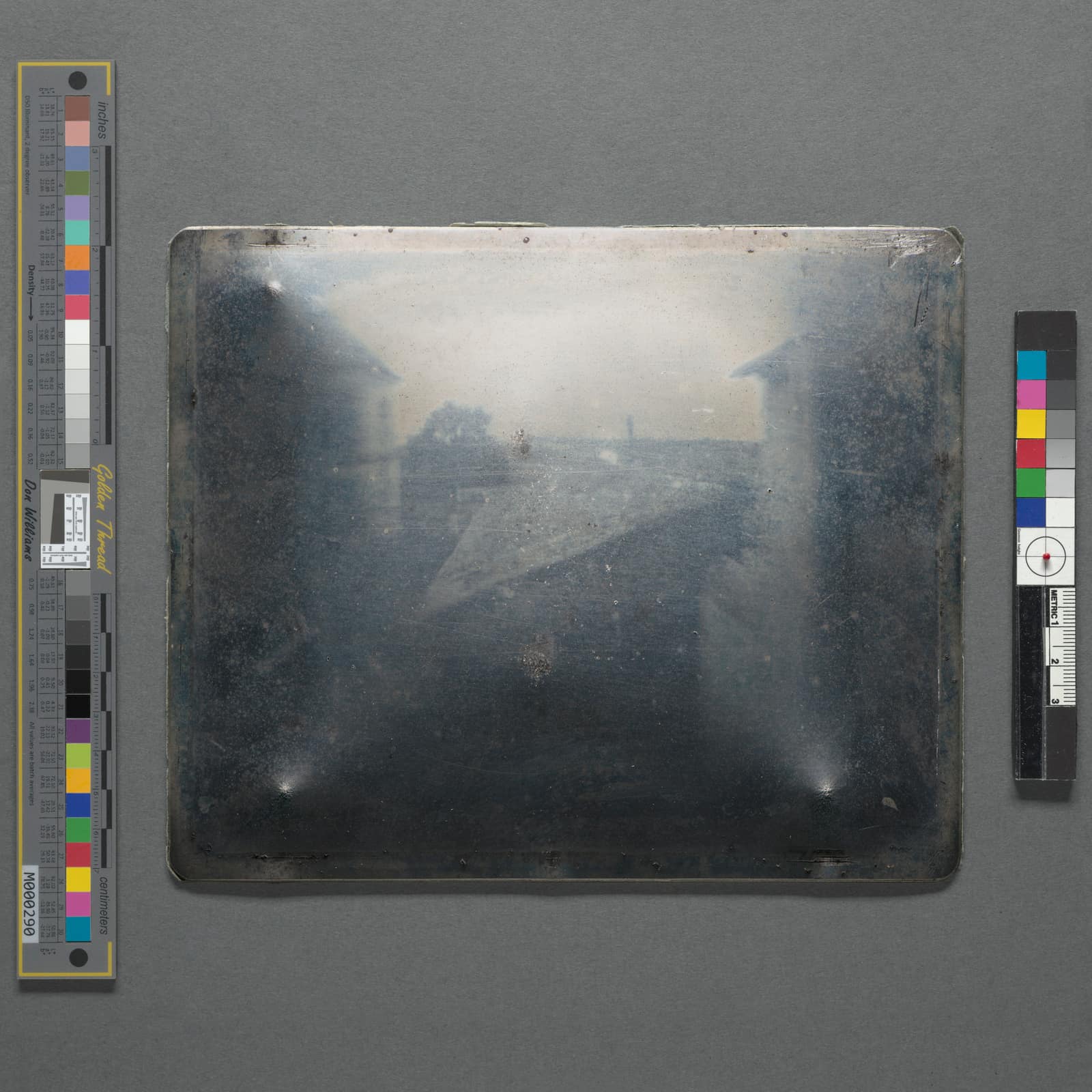 Une plaque de métal presque carrée aux coins légèrement arrondis, photographiée sur un carton gris entre deux bandes de repères de couleur et de gris. Sa surface polie est sombre, comme un miroir, voilée de buée et de petites taches ; ce n'est qu'en regardant de près qu'y apparaissent des formes pâles : des toits qui descendent vers le centre à gauche et à droite, une bande de ciel plus claire, un arbre indistinct, comme vus à travers le brouillard.