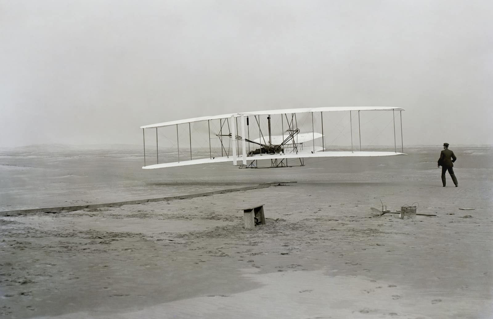 A black and white photograph of a flat, empty stretch of sand under a pale grey sky. Just above the ground, a large biplane of white fabric and thin wooden struts has lifted off, a man lying flat on the lower wing at its centre. To the right, a man in a dark suit and cap stands watching, his arms slightly out from his body; on the sand below the aircraft lie a long wooden launching rail, a small bench and scattered tools.