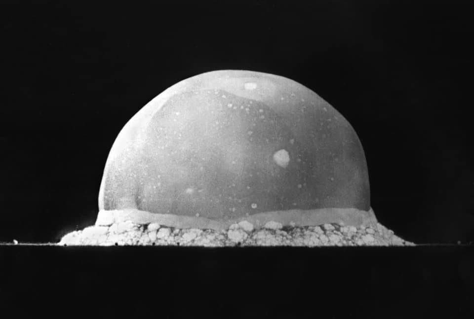 Black and white photograph against a black sky: a pale dome of fire, wider than it is tall, its surface mottled with bright spots, sits on a flat dark horizon; along its base a ragged white rim of dust and debris is being thrown up from the ground.