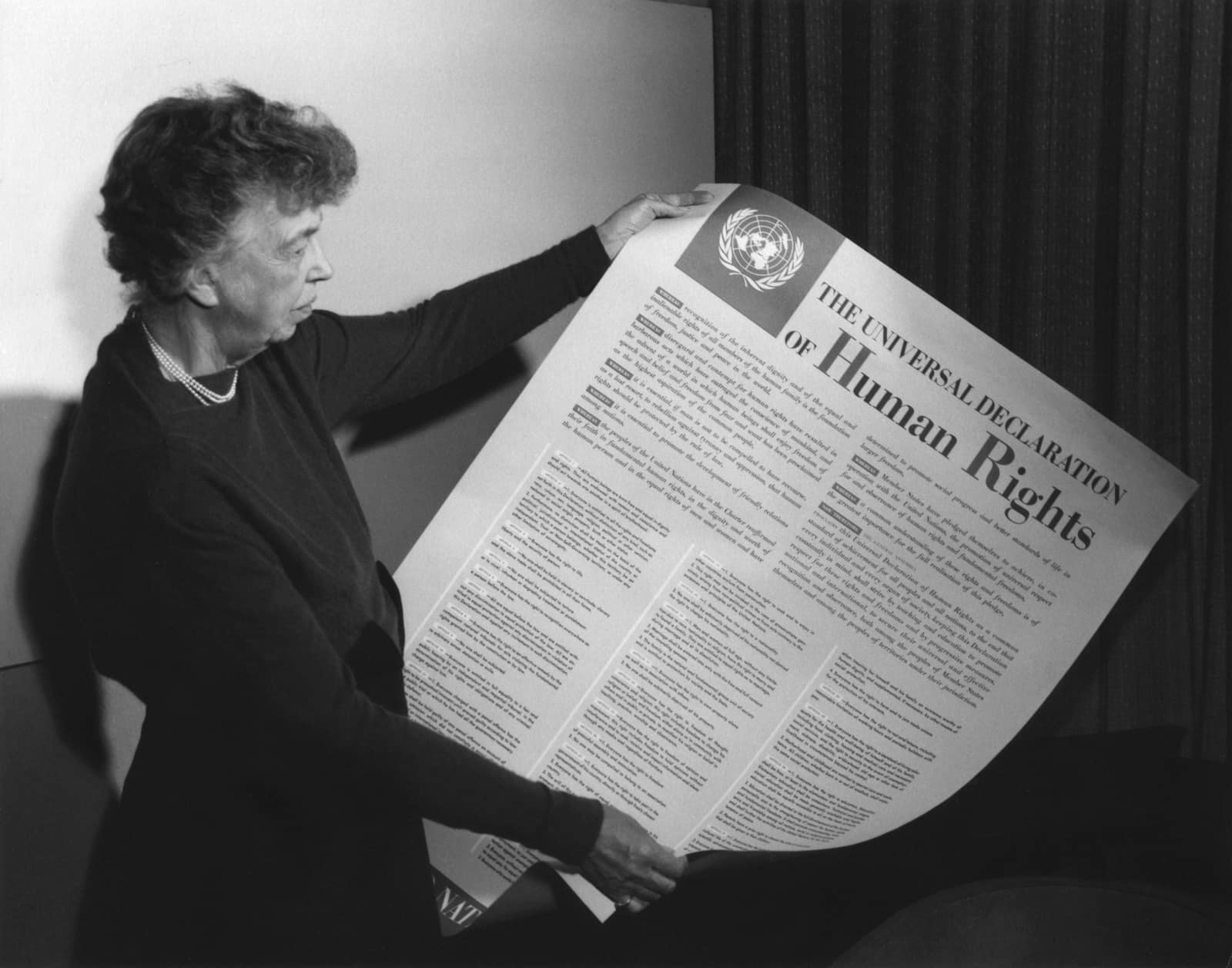 Photographie en noir et blanc de 1949 : une femme aux cheveux courts et ondulés, en robe sombre et collier de perles, se tient de profil et déploie une grande affiche intitulée The Universal Declaration of Human Rights, surmontée de l'emblème des Nations unies et couverte de colonnes en petits caractères.