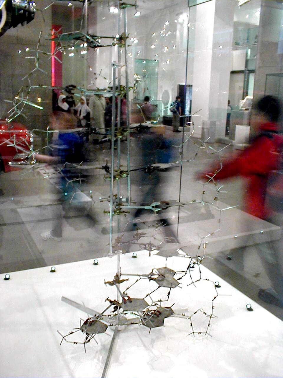 Derrière une vitre, dans une salle de musée, une haute maquette grêle faite de fines tiges de métal et de plaques métalliques plates découpées en forme d'anneaux, empilées autour d'une tige centrale verticale et s'enroulant lentement vers le haut ; à l'arrière-plan passent des visiteurs flous.