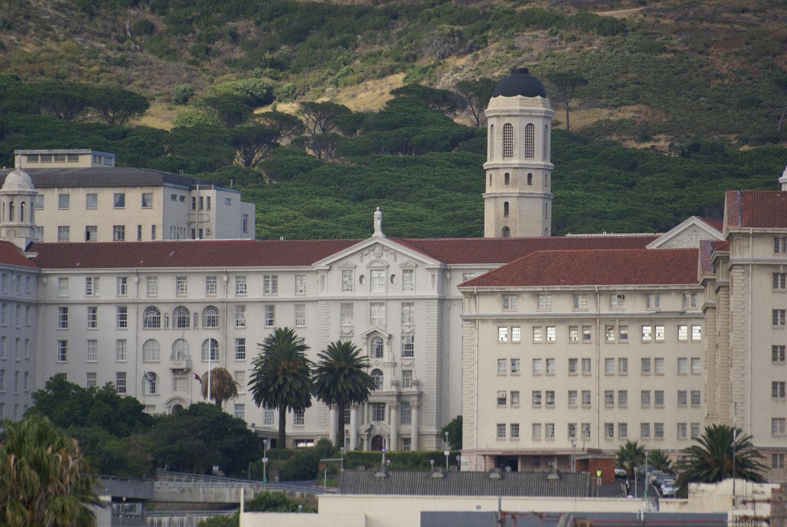 A long pale hospital building of several storeys with red tiled roofs, rows of regular windows and an ornate central gable, flanked by palm trees; a domed bell tower rises behind it against a steep green hillside of pines.
