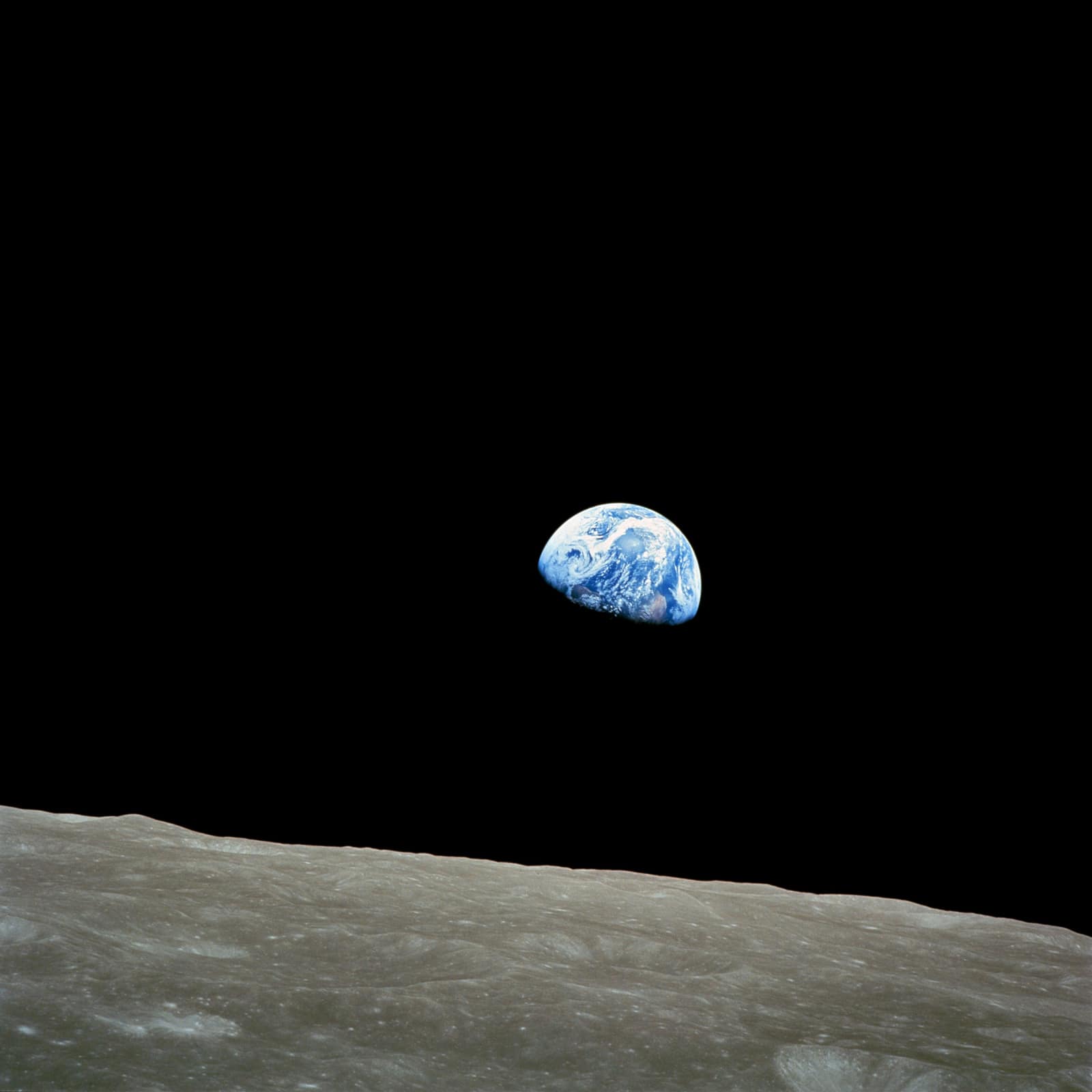 Photographie en couleurs : au-dessus d'un horizon lunaire gris et criblé de cratères, au premier plan, une Terre à demi éclairée flotte dans un ciel entièrement noir, océans bleus striés de nuages blancs, sa moitié inférieure perdue dans l'ombre.