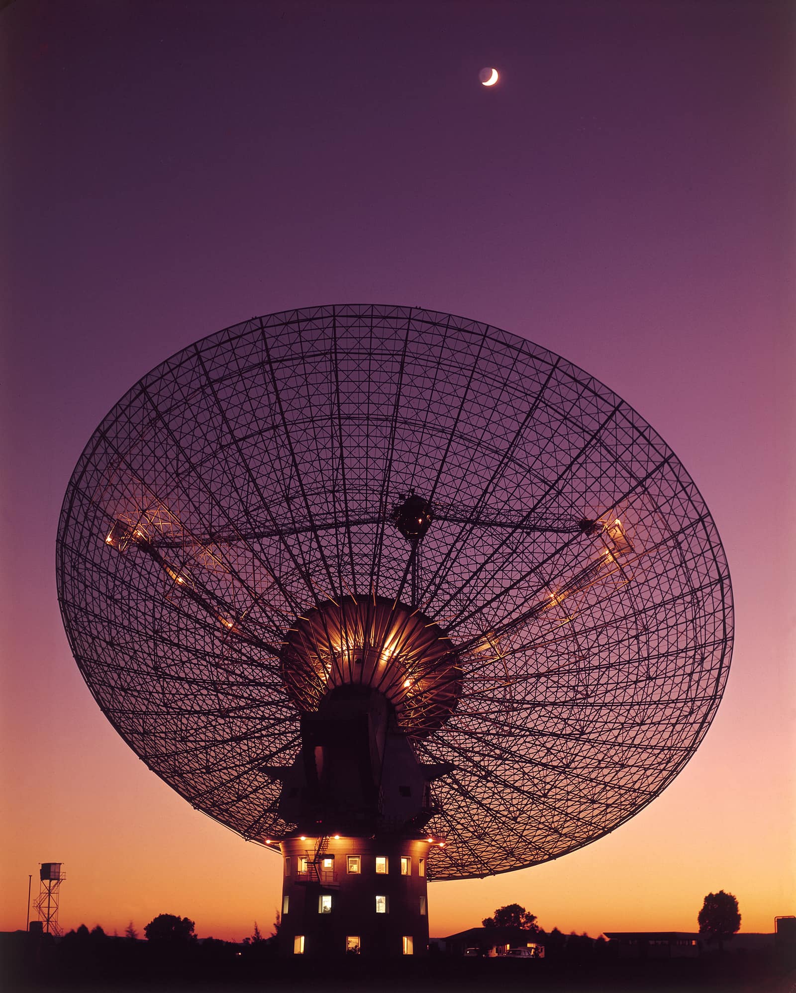 Colour photograph from 1969 at dusk: a huge radio dish, its open steel lattice seen from behind, tilted up on a squat tower with lit windows, silhouetted against a sky fading from orange at the horizon to violet; a thin crescent Moon hangs high above it.