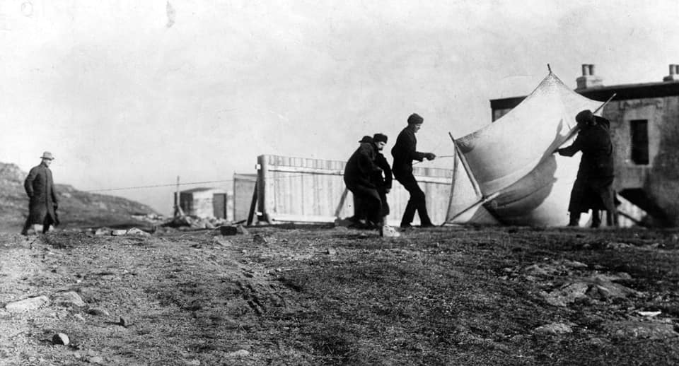 Une photographie en noir et blanc de décembre 1901, sur le sol nu et pierreux de Signal Hill : quatre hommes en manteaux et bonnets sombres peinent à lever contre le vent un grand cerf-volant clair, près d'un bâtiment de pierre à cheminées, tandis qu'à gauche un homme en chapeau et long manteau les observe, devant une palissade de bois.