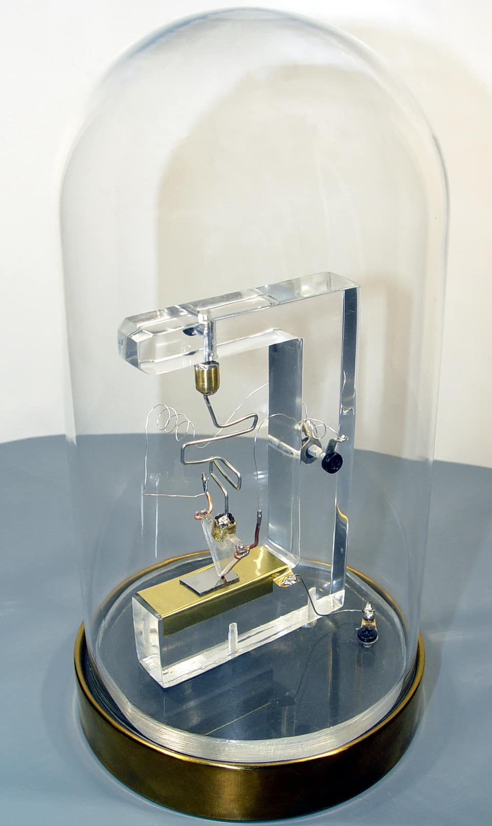 Une réplique du premier transistor sous une haute cloche de verre posée sur un socle de laiton : un cadre en plastique transparent en forme de L renversé maintient un fil fin et coudé qui appuie, par l'intermédiaire d'un petit coin en plastique, sur une plaquette de métal posée sur un bloc doré, entourée de fils de connexion enroulés.