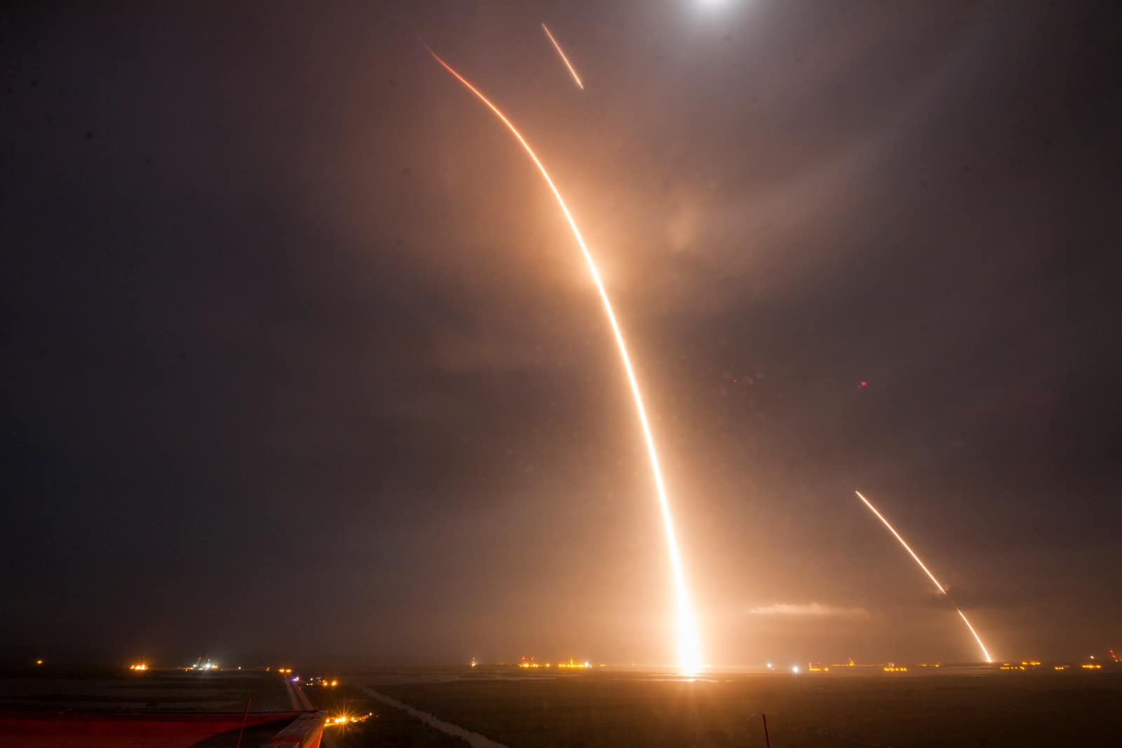 Long exposure photograph at night over flat coastal ground: a bright orange streak rises from the horizon in a tall curve and leaves the frame at the top; a shorter streak high in the sky and a third one on the right, falling back towards the ground, trace the returning first stage's burns; small lights dot the horizon under a hazy, starless sky.