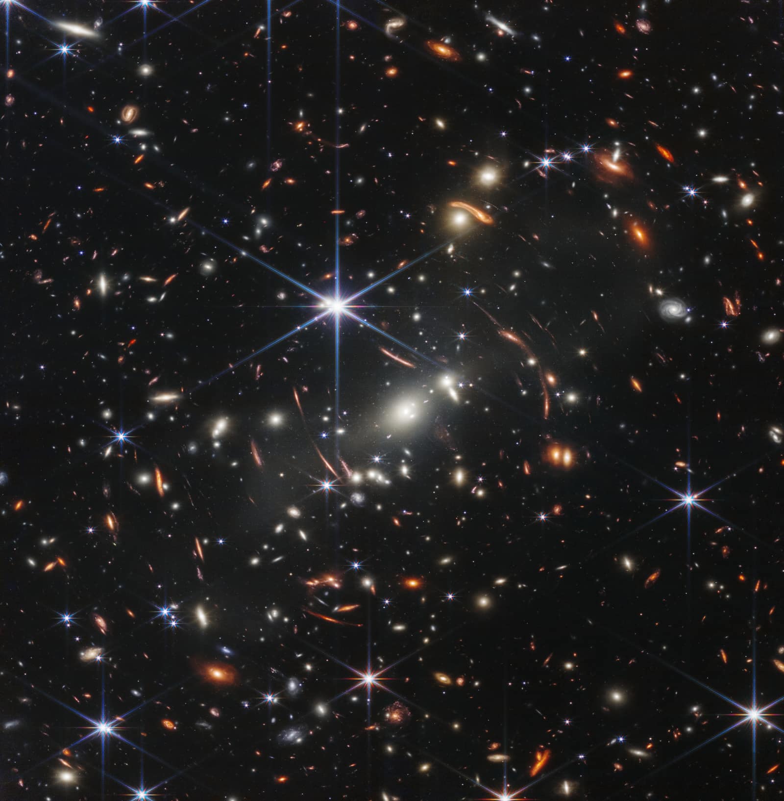 A square of black sky crowded with thousands of small glowing shapes: white, yellow and orange galaxies, some round, some spiral, some stretched into thin curved arcs around a pale cluster of bright galaxies near the centre; a few stars in the foreground stand out with six long spikes of light.