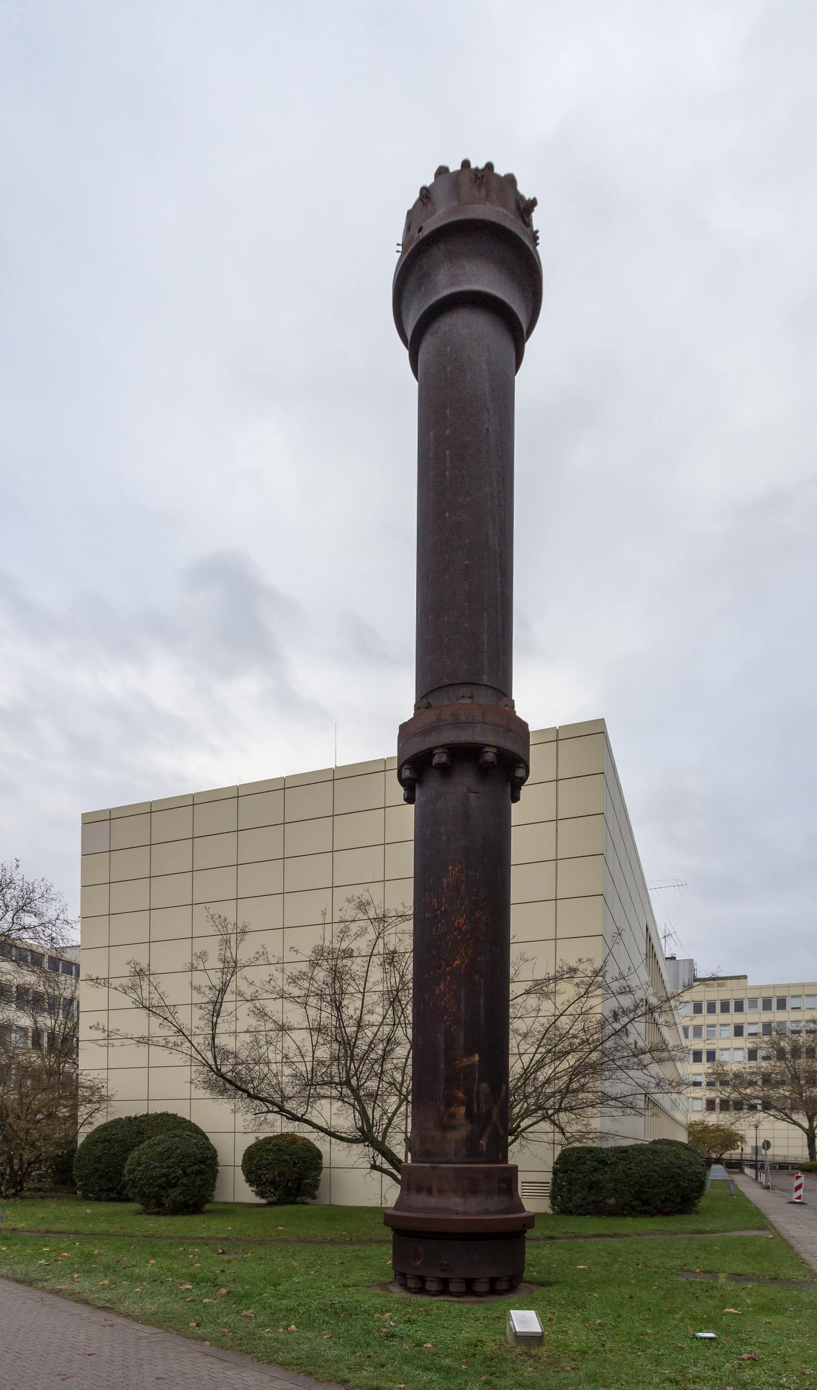 Une haute colonne étroite d'acier sombre et rouillé se dresse sur une pelouse, bien plus haute que les buissons qui l'entourent, sous un ciel gris et couvert. Elle s'épaissit au sommet en un lourd collier couronné de boulons, et à mi-hauteur comme à sa base en brides boulonnées. Derrière elle s'élève la façade claire, carrelée, d'un bâtiment universitaire moderne.